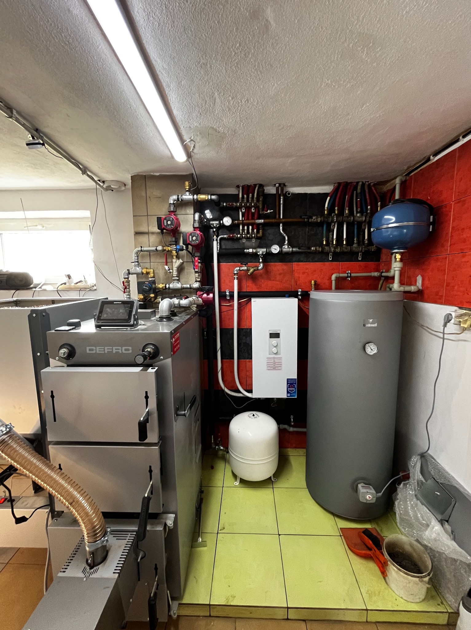 Nowoczesna kotłownia z piecem Defro, zbiornikiem Galmet i rozbudowaną instalacją hydrauliczną na tle czerwono-czarnych płytek. Widoczne rury i osprzęt.
