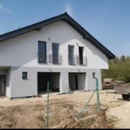 Nowoczesny dom jednorodzinny w Wejherowie z szarą elewacją, czarnymi ramami okiennymi i balkonami, w trakcie wykończenia. Widoczne przygotowania do zagospodarowania terenu wokół budynku.