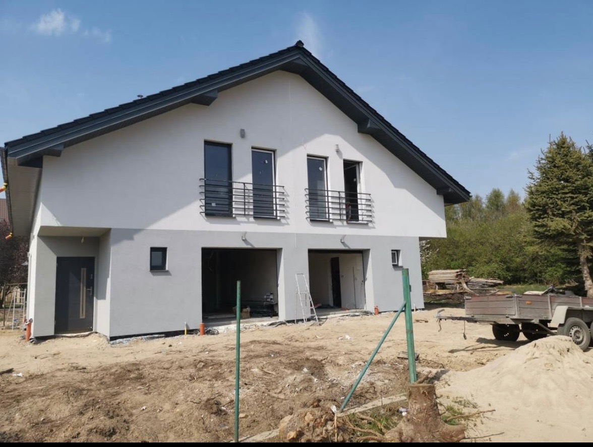 Nowoczesny dom jednorodzinny w Wejherowie z szarą elewacją, czarnymi ramami okiennymi i balkonami, w trakcie wykończenia. Widoczne przygotowania do zagospodarowania terenu wokół budynku.