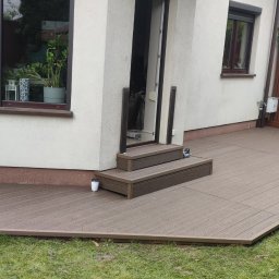 Volodymyr Dik - Nowoczesny taras z deski kompozytowej w kolorze brązowym, przylegający do budynku z dwoma stopniami wejściowymi. Widoczny fragment ogrodu i drewniane ogrodzenie.