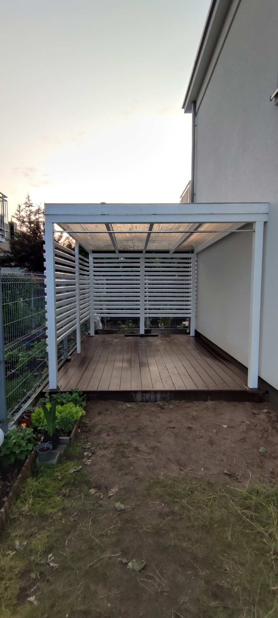 Nowoczesna, biała pergola z drewnianą podłogą tarasową, przylegająca do budynku. Widoczne poziome lamele i przezroczysty dach. Otoczenie w trakcie aranżacji.