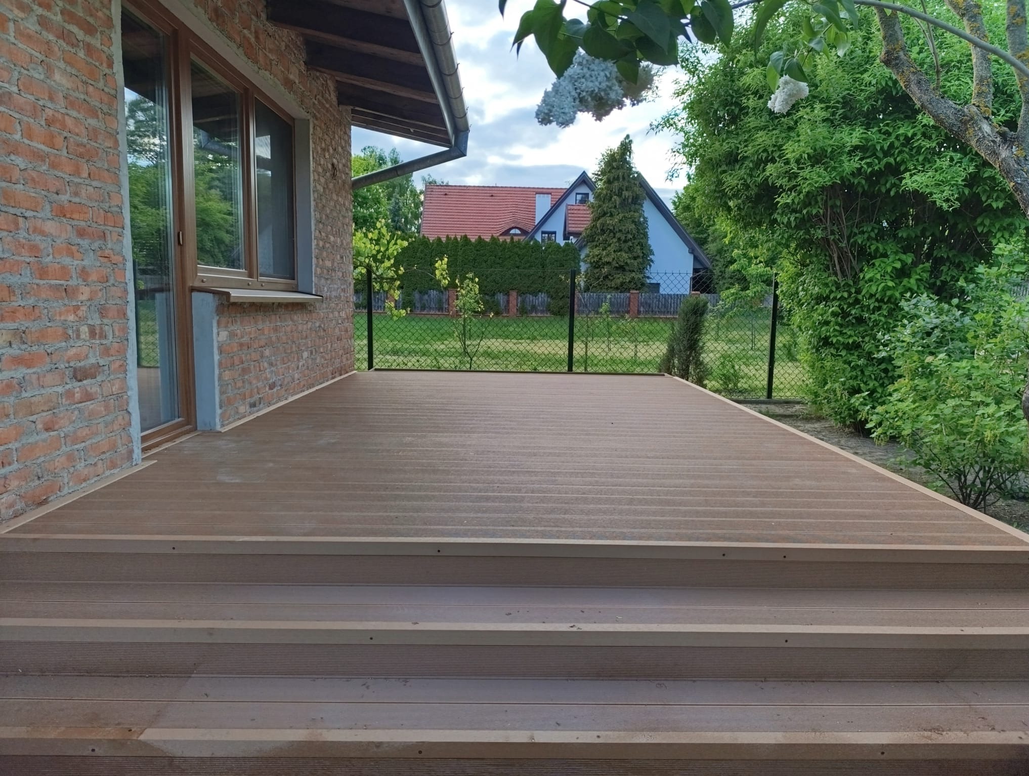 Nowoczesny taras kompozytowy z trzema stopniami, przylegający do ceglanego budynku. Widok na ogród z siatką ogrodzeniową i dom w tle.