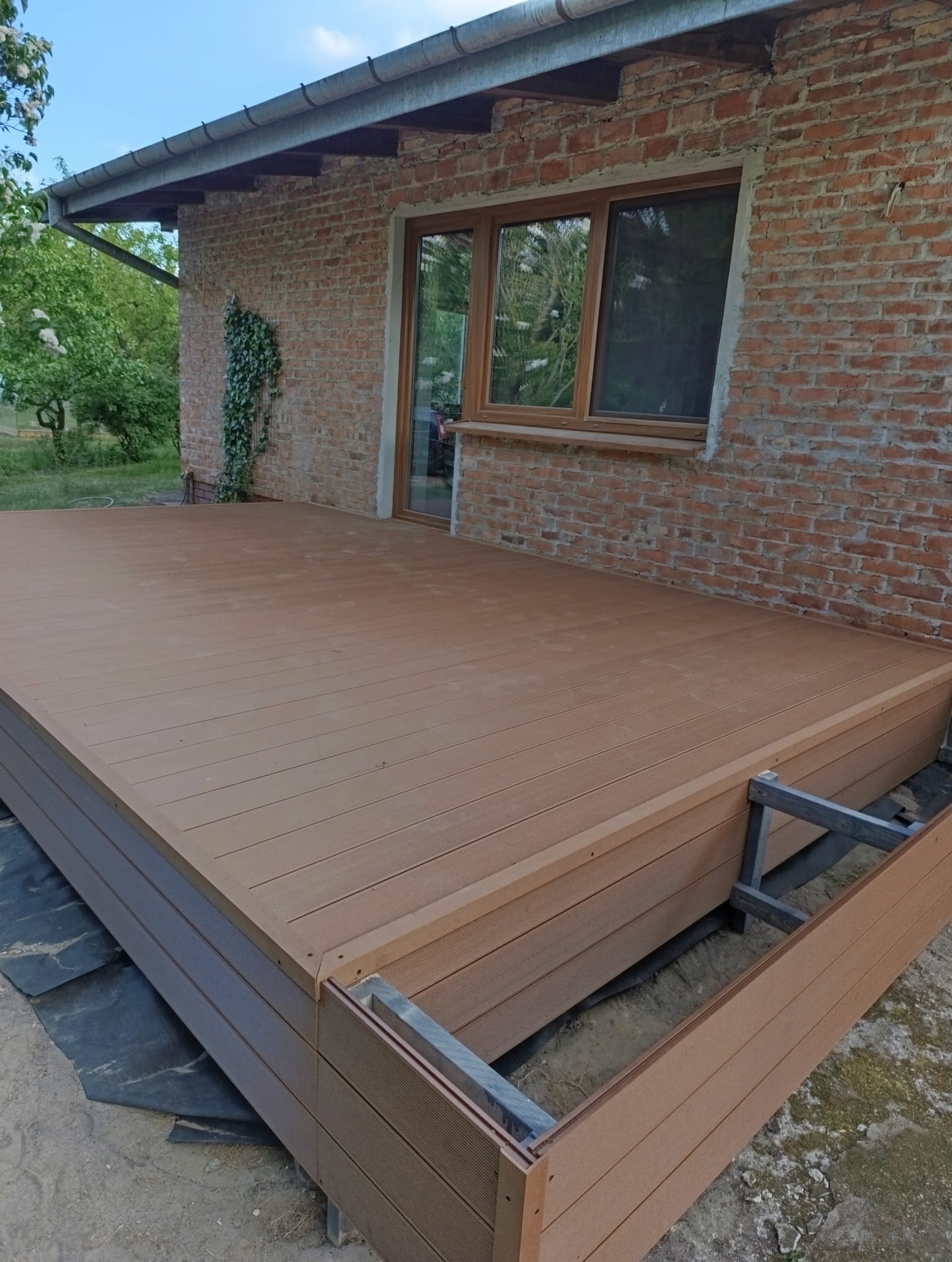 Brązowy taras kompozytowy przy ceglanym budynku, widoczne elementy konstrukcyjne i podłoże. Ujęcie z niskiej perspektywy podkreśla detale montażu.