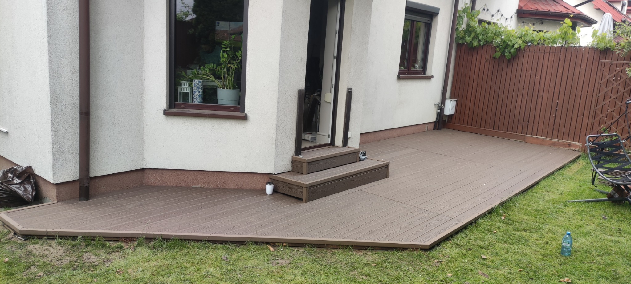 Nowoczesny taras z deski kompozytowej w kolorze brązowym, przylegający do budynku z dwoma stopniami wejściowymi. Widoczny fragment ogrodu i drewniane ogrodzenie.