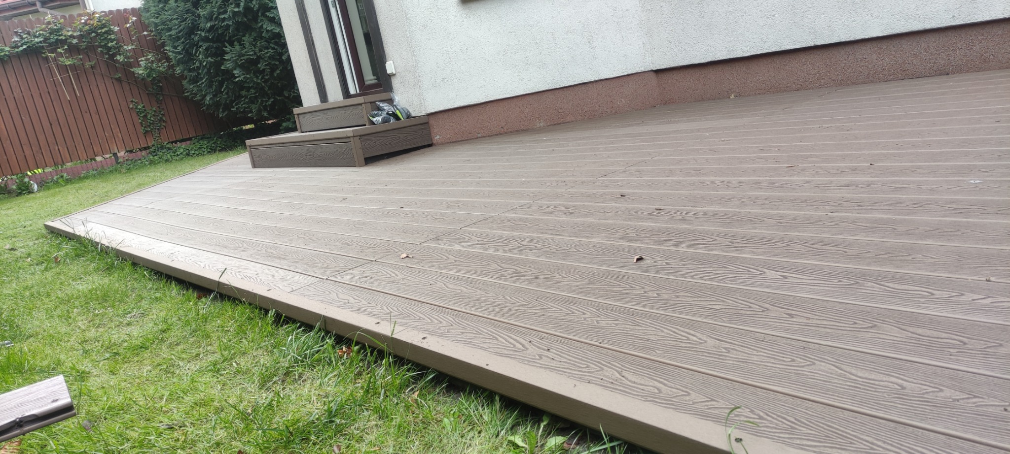 Beżowy taras kompozytowy przylegający do budynku, z widocznymi schodkami wejściowymi i fragmentem trawnika. Ujęcie z niskiej perspektywy podkreśla strukturę desek.