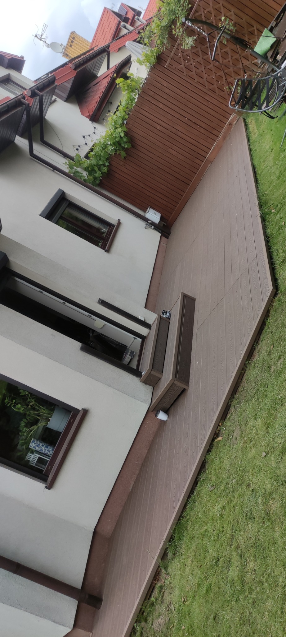 Nowoczesny taras z deski kompozytowej w odcieniach brązu, przylegający do jasnego budynku z brązowymi oknami. Widoczne schody i fragment trawnika. Ujęcie z dołu.