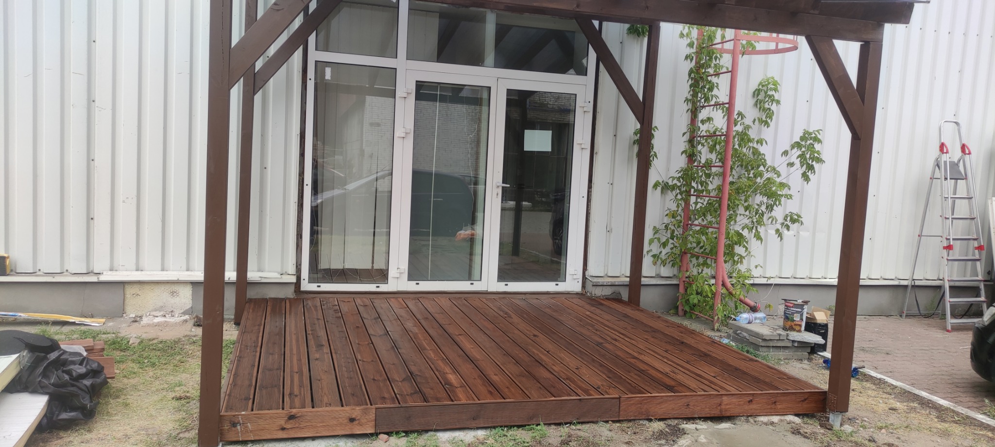 Drewniany taras zadaszony pergolą, brązowe deski, białe drzwi balkonowe, drabina i pnącza w tle. Widok na fragment posesji z elementami budowlanymi.