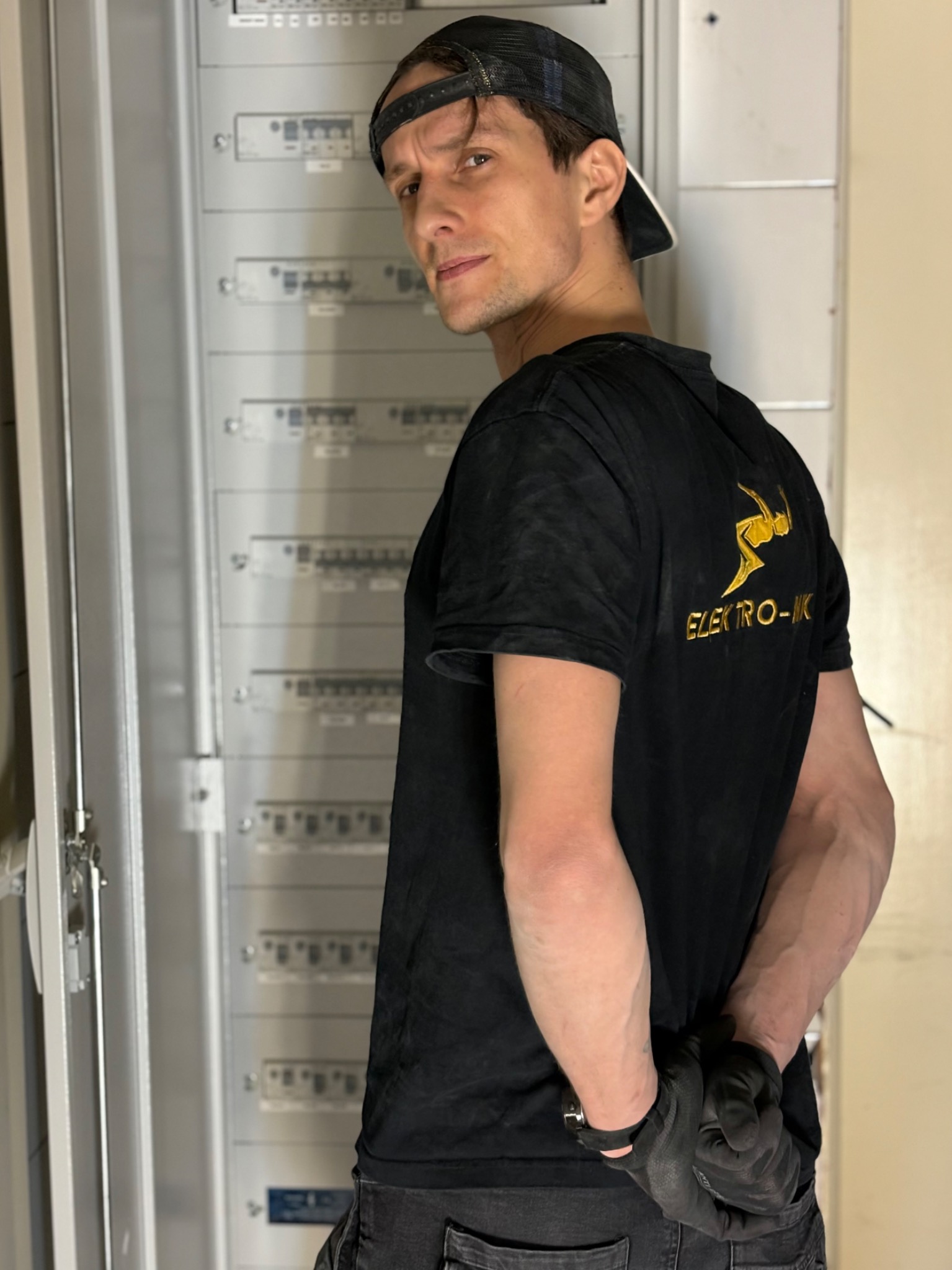 Elektryk w czarnej koszulce z logo firmy, stojący przed otwartą skrzynką bezpiecznikową, z rękami założonymi za plecami i czarnymi rękawicami roboczymi.