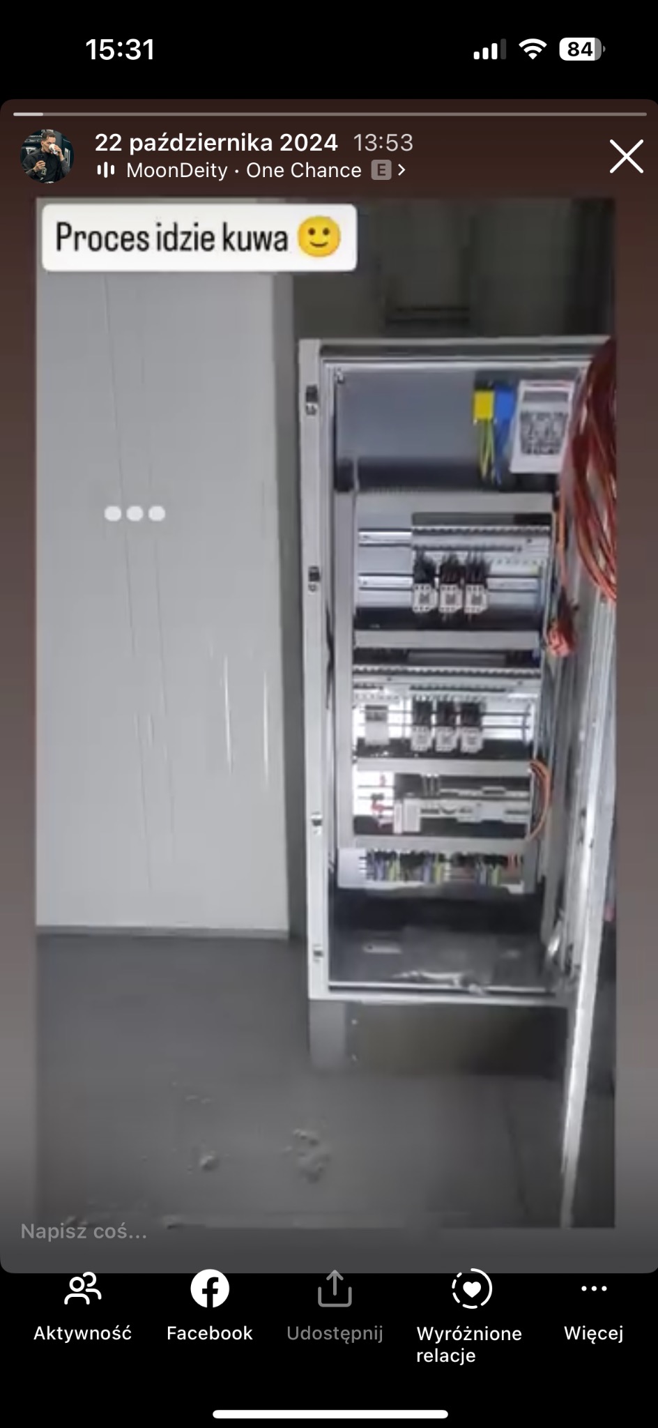 Otwarta szafa elektryczna z widocznymi elementami instalacji, takimi jak bezpieczniki i przewody, umieszczona w surowym pomieszczeniu o szarych ścianach i podłodze.