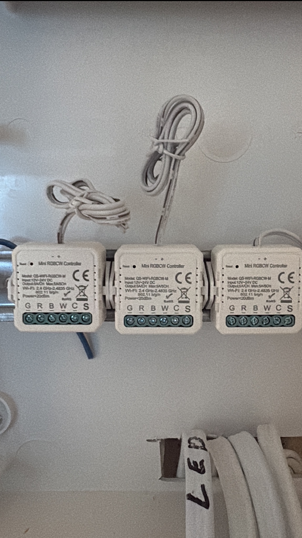 Trzy białe kontrolery LED RGBW zamontowane na szynie DIN w białej skrzynce, z widocznymi przewodami i oznaczeniami. Kontrolery marki Mini RGBCW Controller.