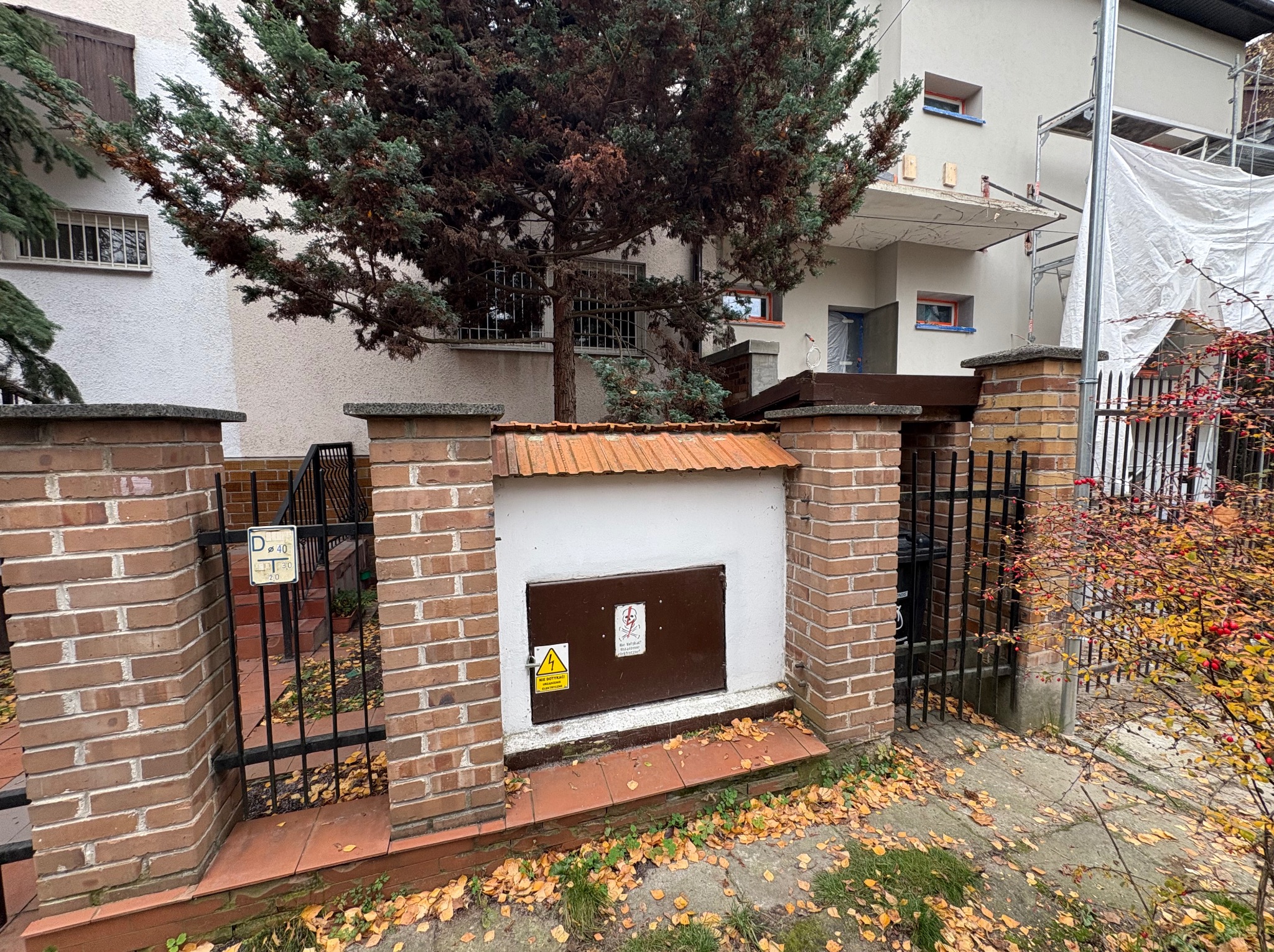 Zamurowana skrzynka elektryczna z brązowymi drzwiczkami i ostrzeżeniami, otoczona ceglanym murem i metalową bramką, na tle budynku w trakcie remontu.