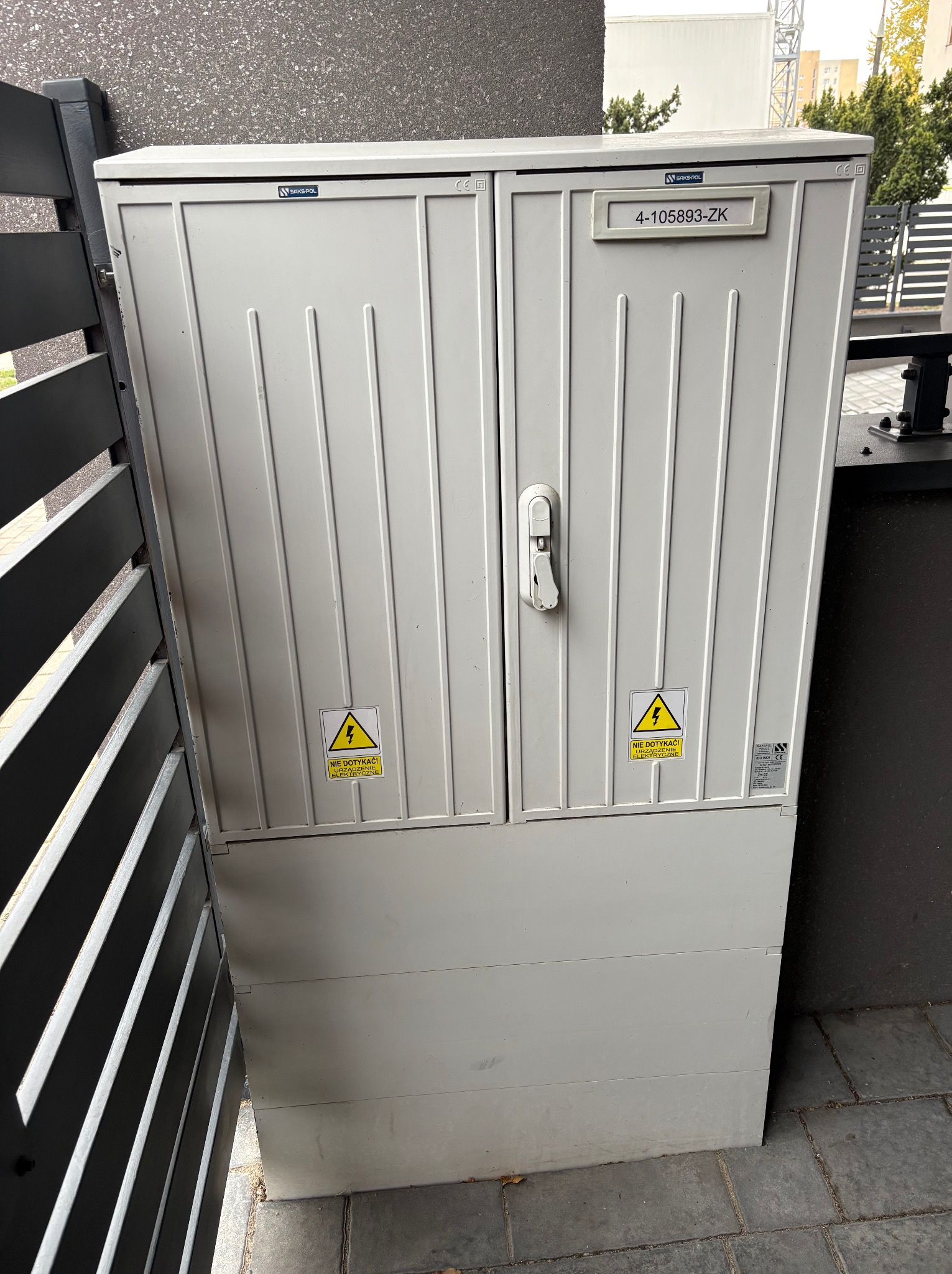 Szara szafka elektryczna z ostrzeżeniami 'Nie dotykać! Urządzenie elektryczne' na zewnątrz, umieszczona przy szarym murze i płocie. Widoczny numer identyfikacyjny.