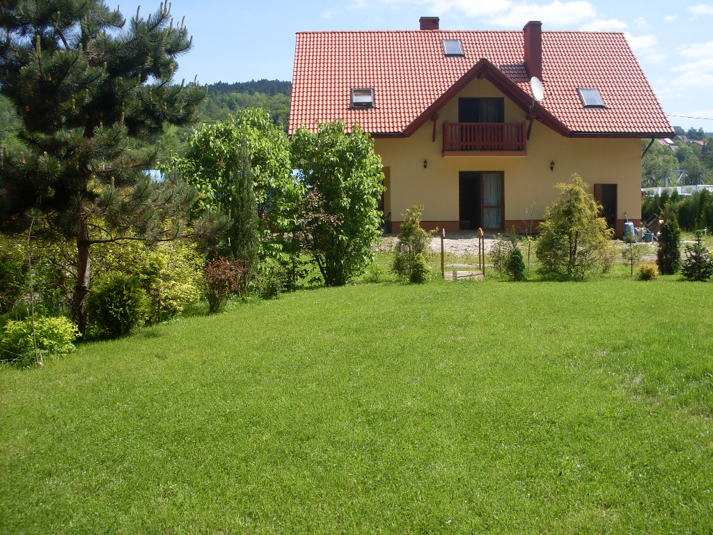 Żółty dom z czerwonym dachem i balkonem, otoczony zielenią i trawnikiem, widok z poziomu trawy.