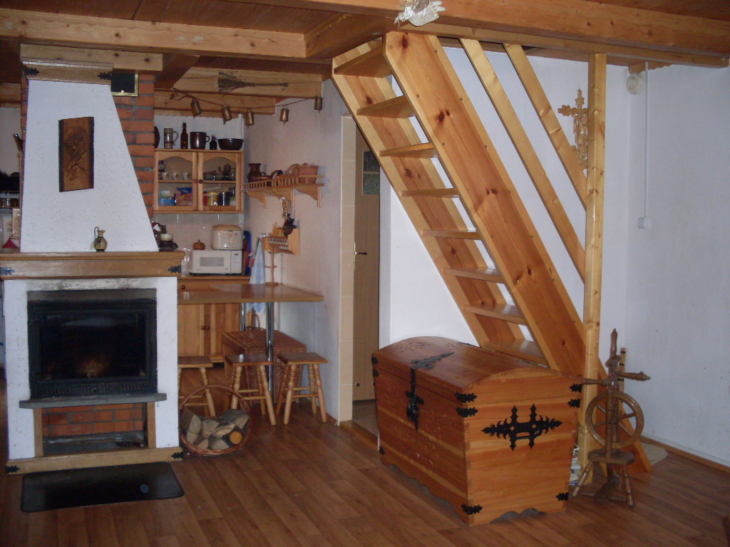 Drewniany domek letniskowy: wnętrze z kominkiem, schodami i skrzynią w rustykalnym stylu. Widoczny kosz z drewnem obok kominka.