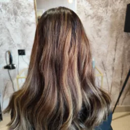 Tył głowy kobiety z brązowymi włosami z blond pasemkami, delikatne fale, widoczne lustro i suszarka w tle.