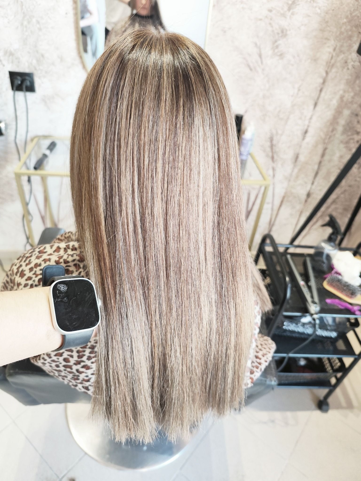 Jasne, proste włosy po koloryzacji widoczne z tyłu głowy klientki w salonie fryzjerskim w Płocku, na pierwszym planie fragment ręki z smartwatchem, w tle wózek fryzjerski z akcesoriami.