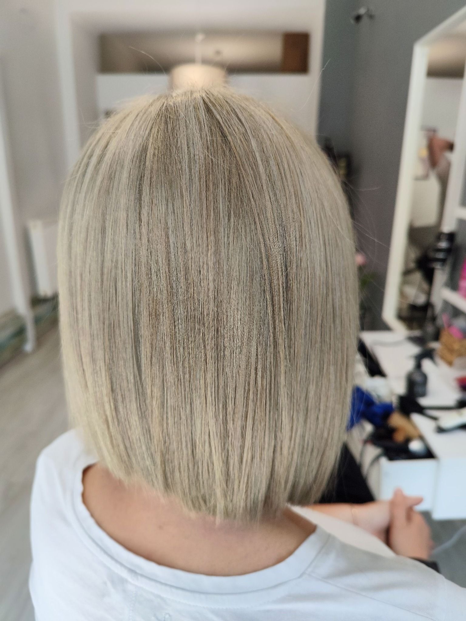 Tył głowy kobiety z krótką, prostą fryzurą w odcieniach blondu; widoczne pojedyncze włosy odstające na czubku głowy, tło rozmyte z elementami wyposażenia salonu.