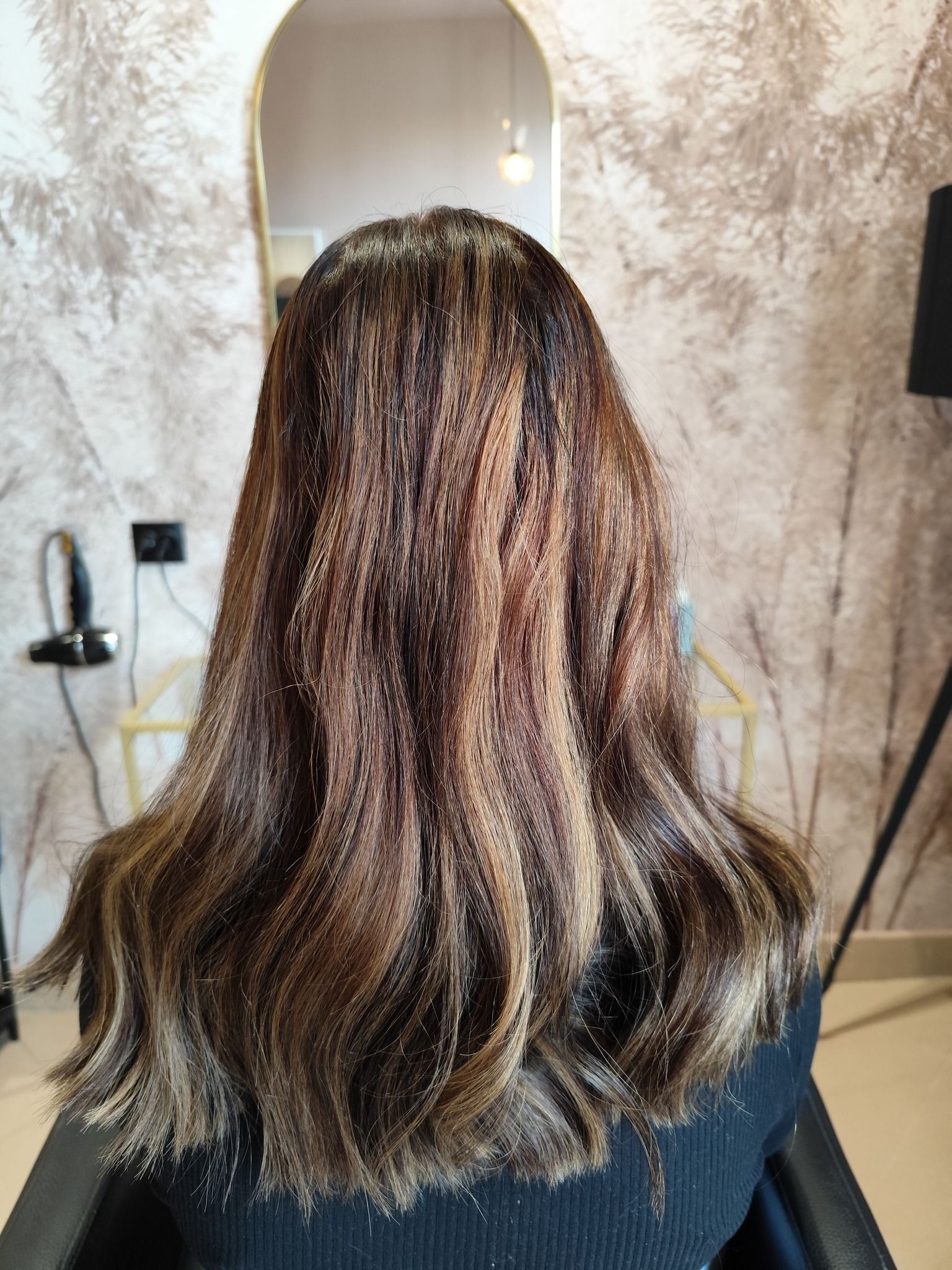 Tył głowy kobiety z brązowymi włosami z blond pasemkami, delikatne fale, widoczne lustro i suszarka w tle.