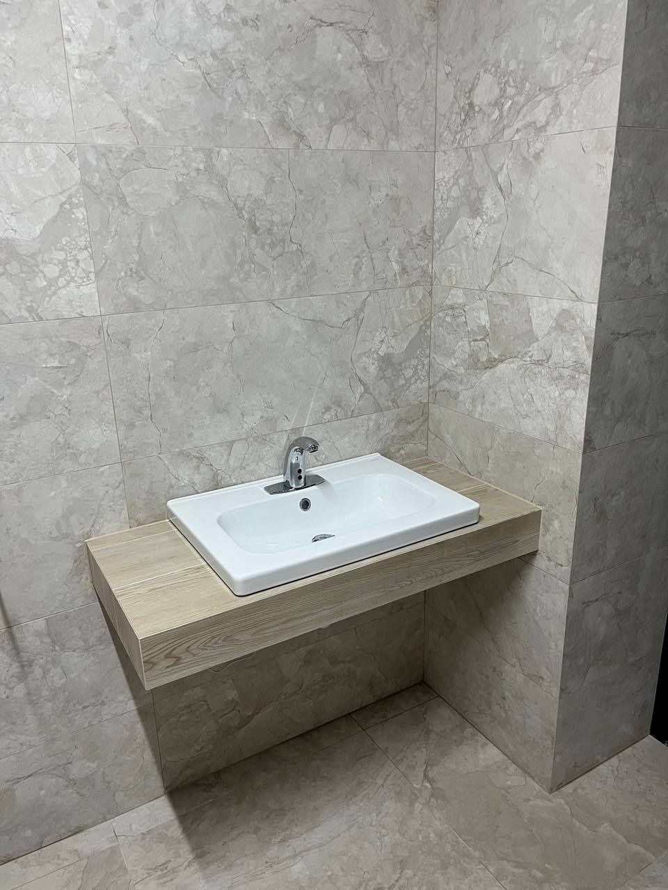 Biała umywalka z chromowaną baterią, zamontowana na drewnopodobnym blacie w łazience wykończonej płytkami imitującymi marmur w odcieniach szarości i beżu.