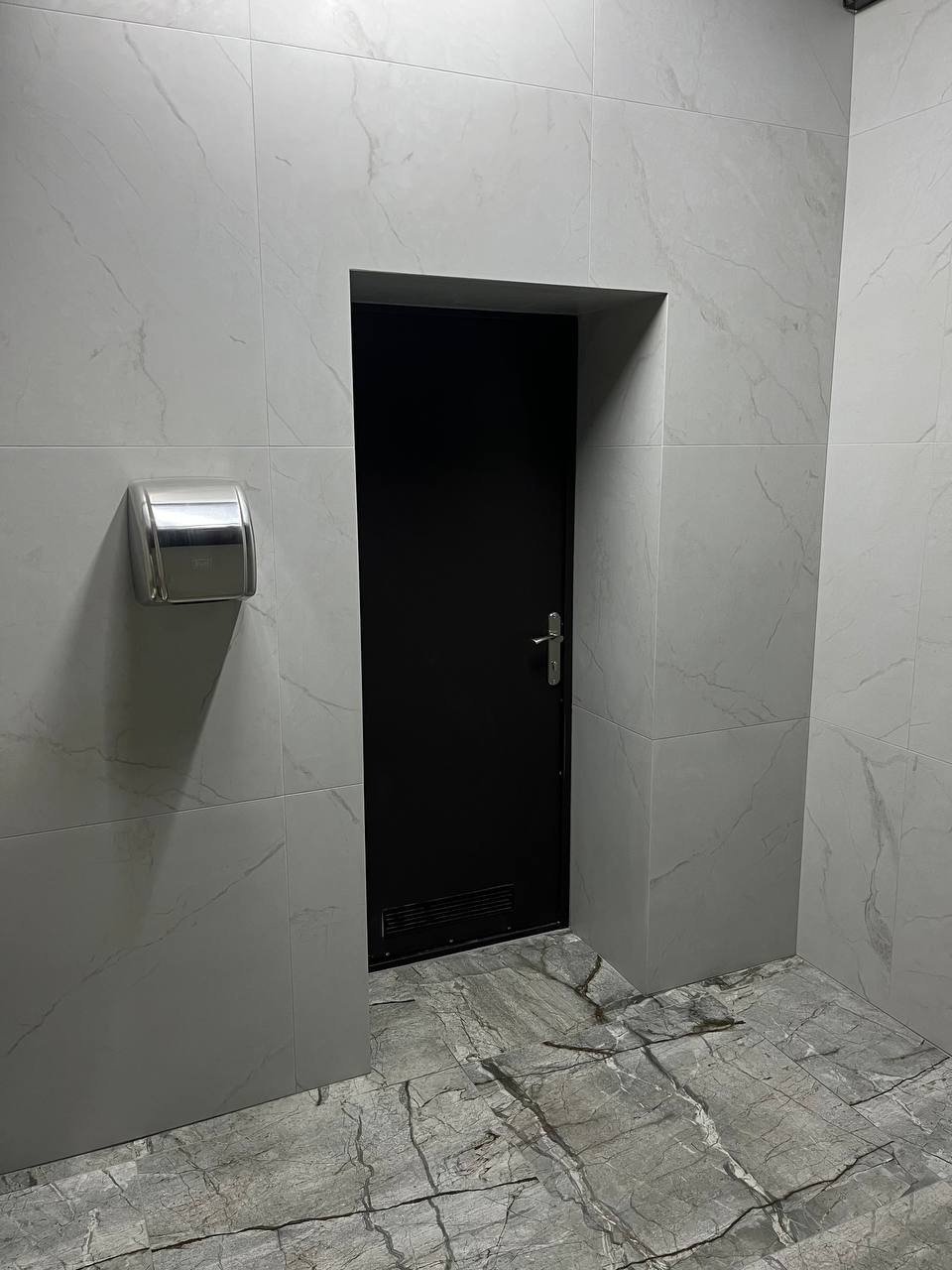 Wykończona łazienka z szarymi płytkami imitującymi marmur na podłodze i ścianach, czarne drzwi z metalową klamką oraz metalowy podajnik na ręczniki papierowe.