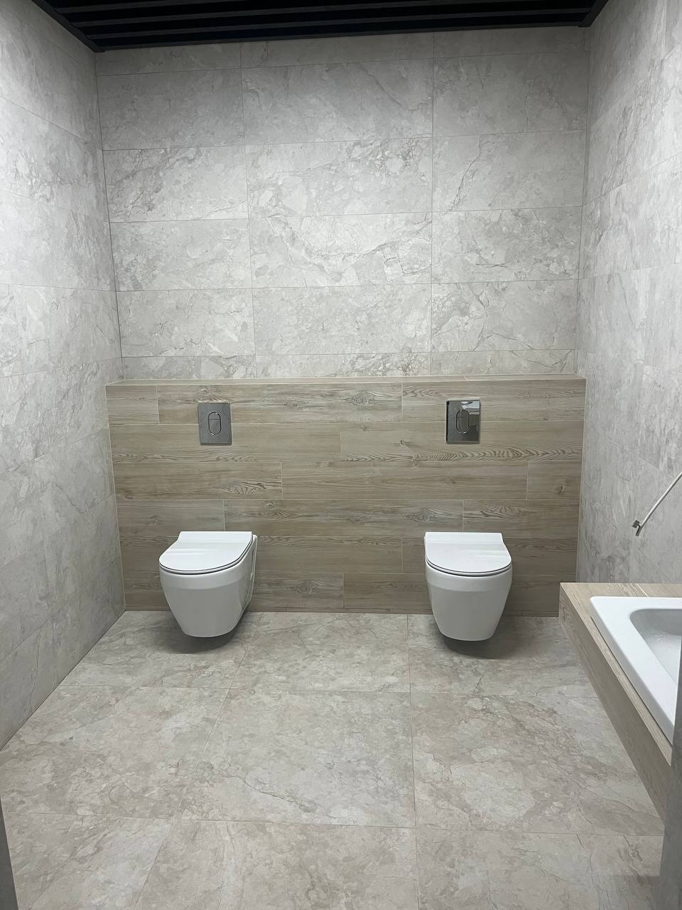 Wykończona łazienka z dwoma podwieszanymi toaletami w jasnych odcieniach szarości i beżu, płytki imitujące marmur na ścianach i podłodze, drewnopodobna zabudowa stelaży podtynkowych.