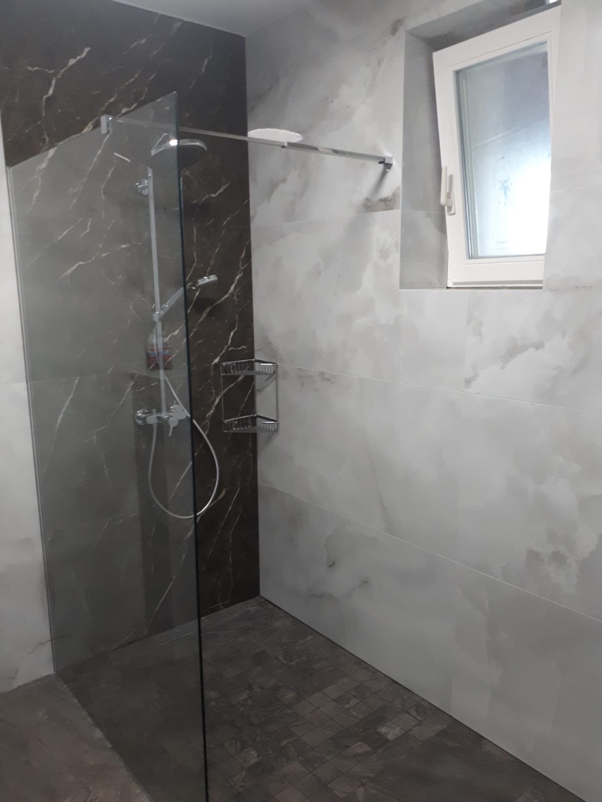 Nowoczesna kabina prysznicowa z marmurowymi płytkami w odcieniach szarości i brązu, widoczny chromowany zestaw prysznicowy i półka na kosmetyki, małe okno z białym obramowaniem w tle.