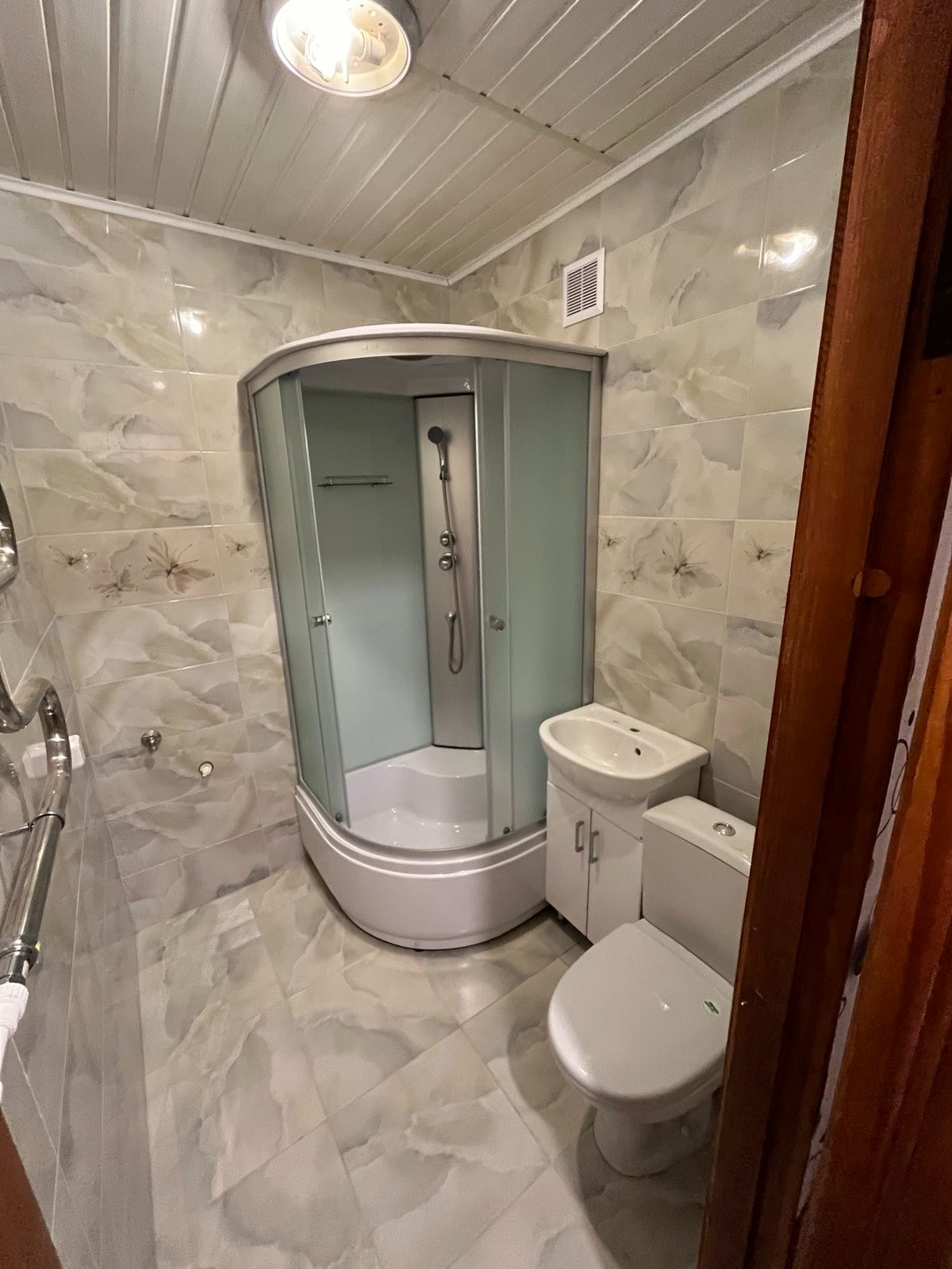 Wykończona łazienka z narożną kabiną prysznicową, umywalką z szafką i toaletą, wyłożona płytkami imitującymi marmur z delikatnym kwiatowym wzorem, oświetlona centralnym plafonem.