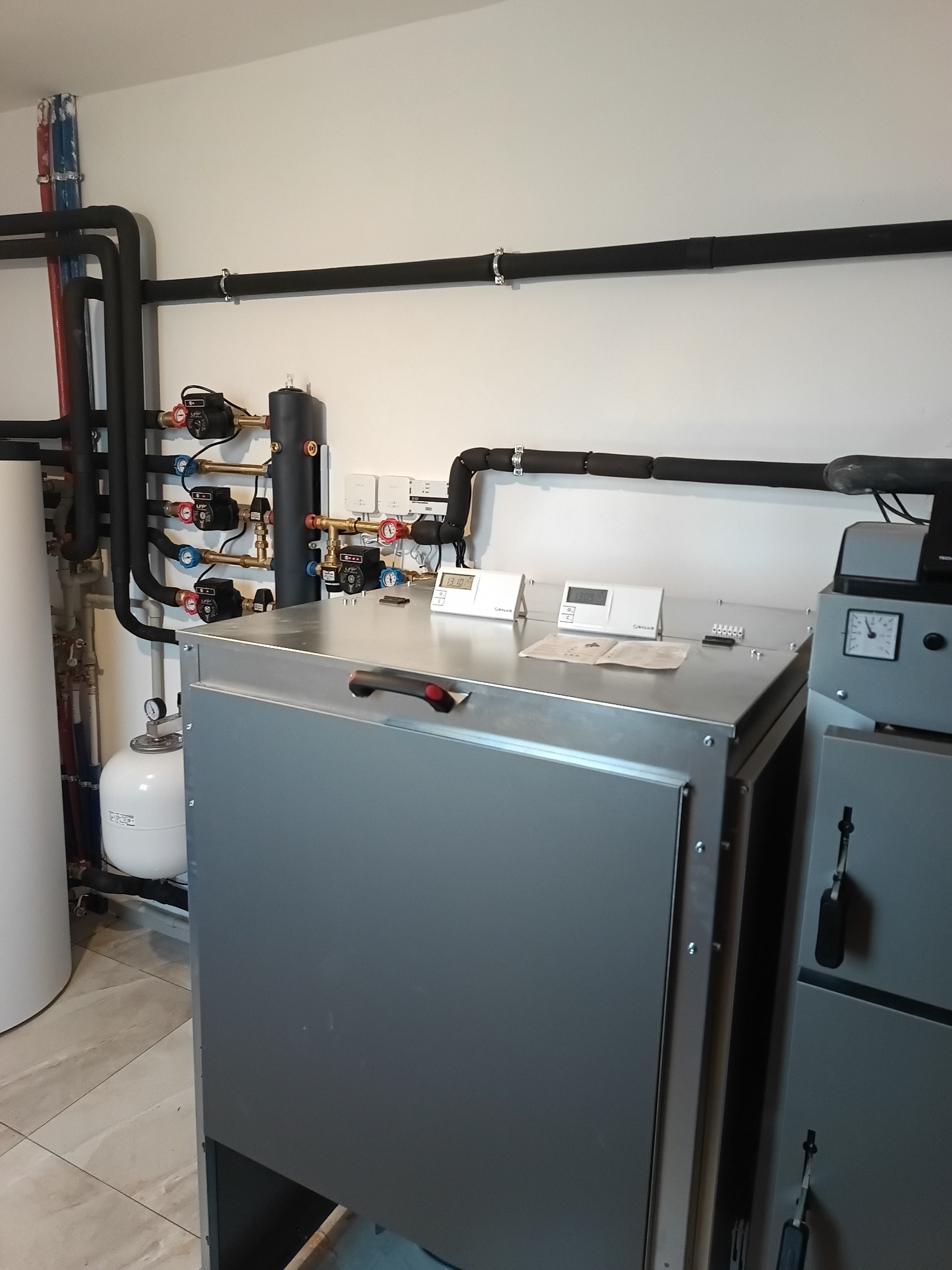 Złożona instalacja grzewcza z kotłem, rurami i zaworami w pomieszczeniu. Widoczne czujniki i manometry. Elementy instalacji są zaizolowane.