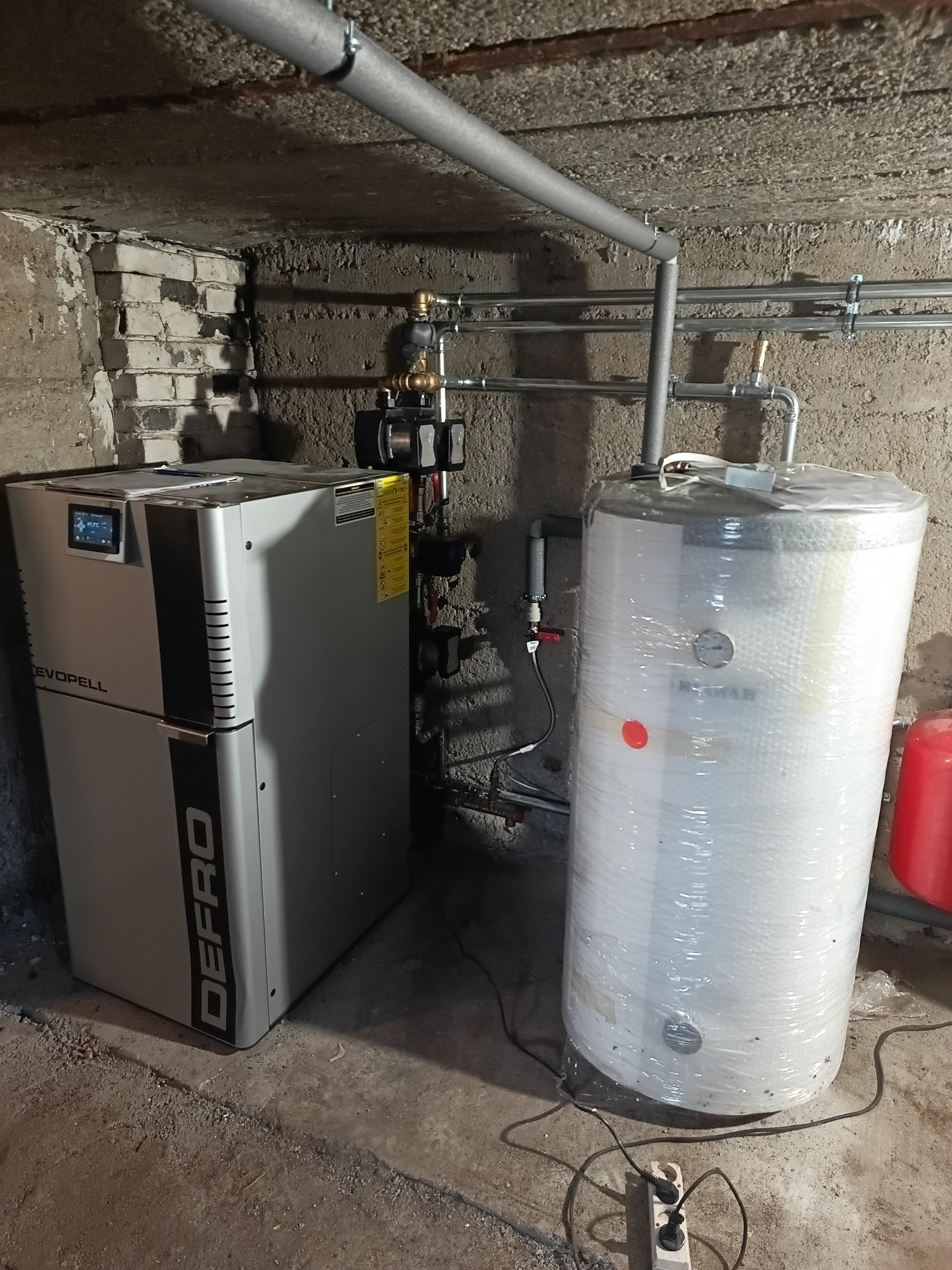 Nowoczesny piec Defro Pell z zasobnikiem ciepłej wody, połączone rurami w kotłowni. Zasobnik owinięty folią ochronną. Widoczne elementy instalacji CO.