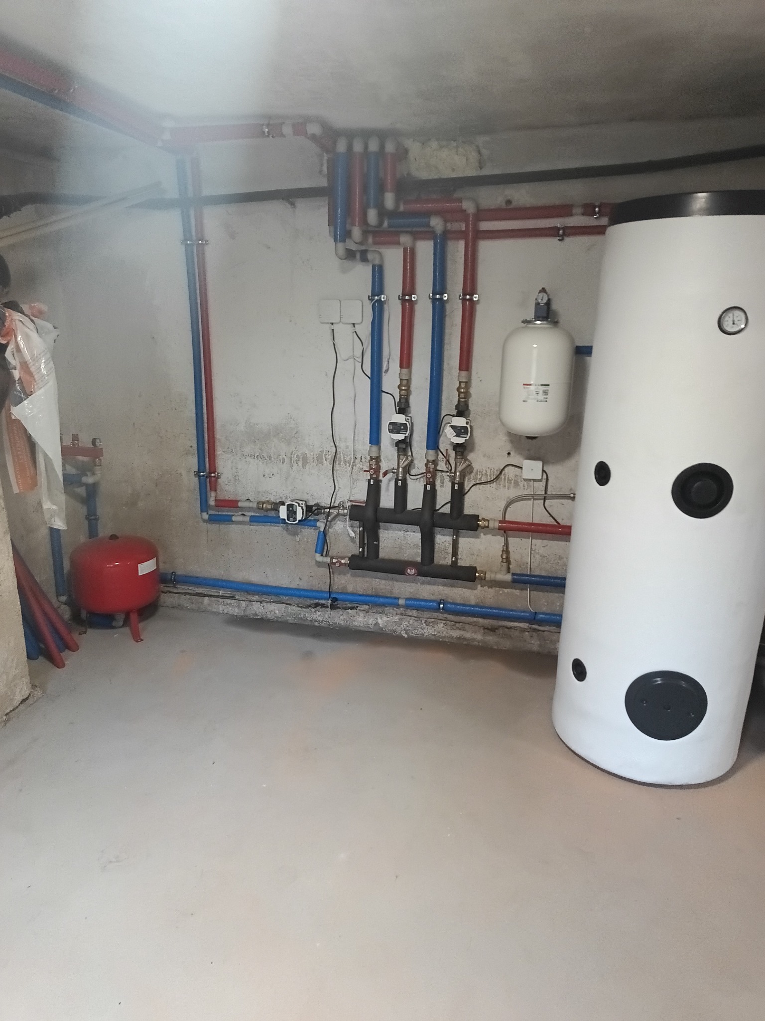 Zakończona instalacja grzewcza w piwnicy: biały zasobnik ciepłej wody, czerwone i niebieskie rury, czarne elementy armatury. Widoczne manometry i zawory.