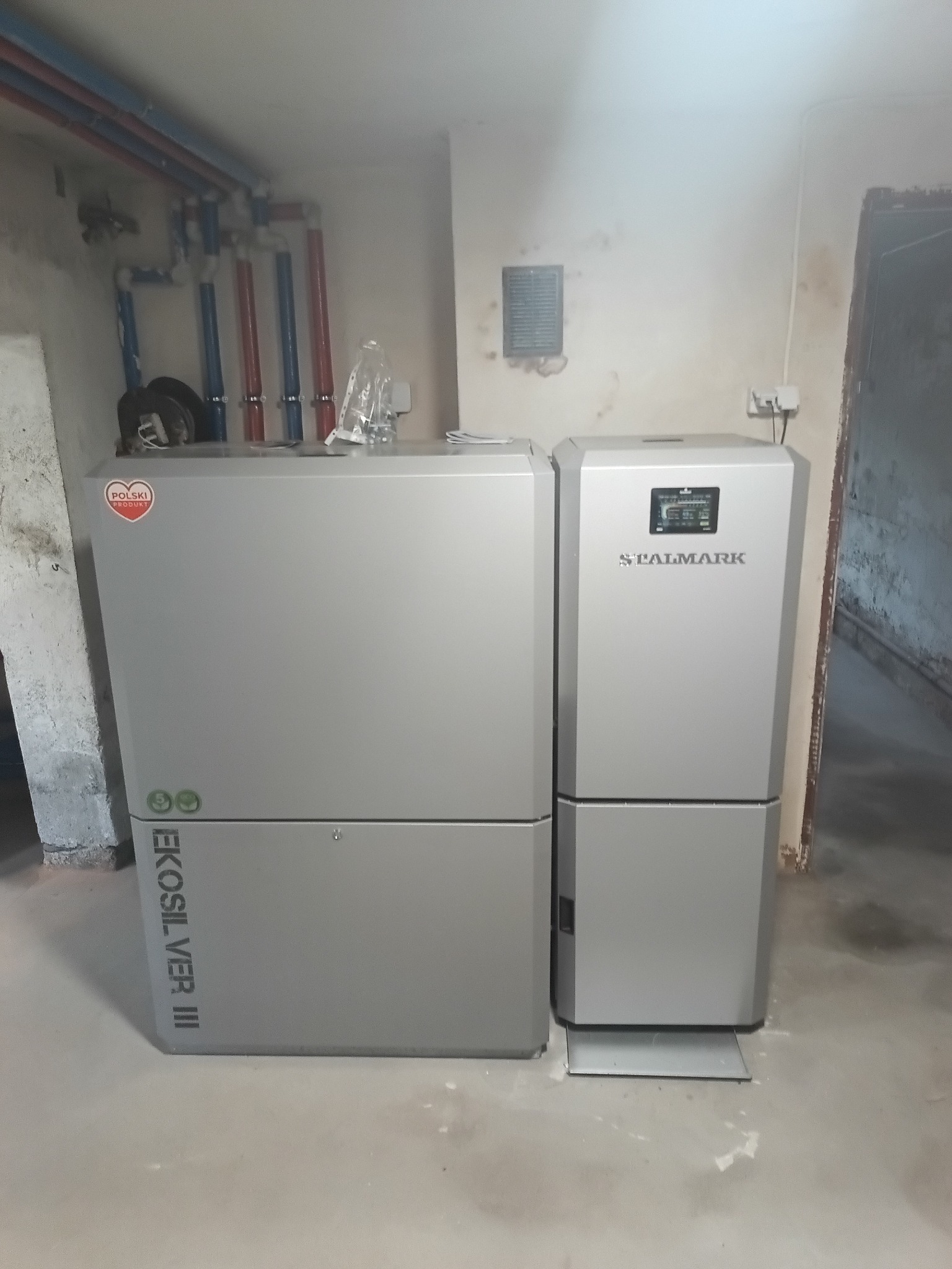 Szary piec CO Stalmark i EkoSilver III w kotłowni z rurami instalacji grzewczej. Widoczne przyłącza i elementy sterujące. Polski produkt.