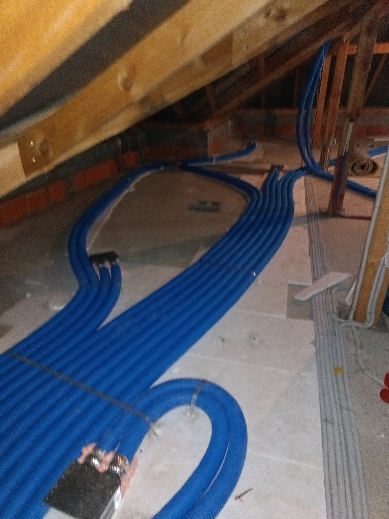 Niebieskie rury wentylacyjne ułożone równolegle na poddaszu, widoczne drewniane elementy konstrukcji dachu i fragmenty instalacji elektrycznej w szarych rurach.