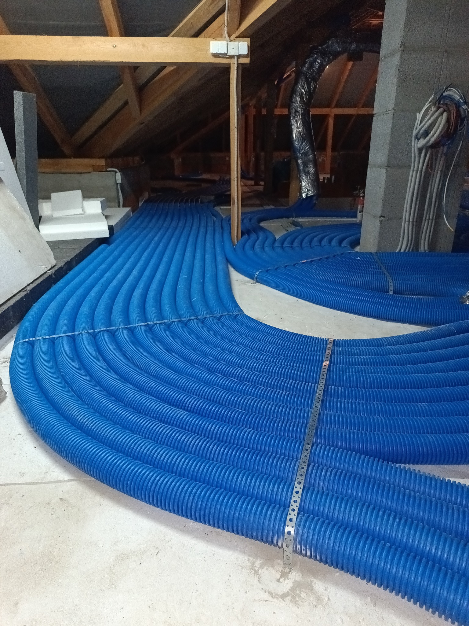 Niebieskie rury rekuperacji ułożone równolegle na poddaszu, widoczne elementy konstrukcji dachu i izolacji. Metalowe mocowania utrzymują rury w porządku.