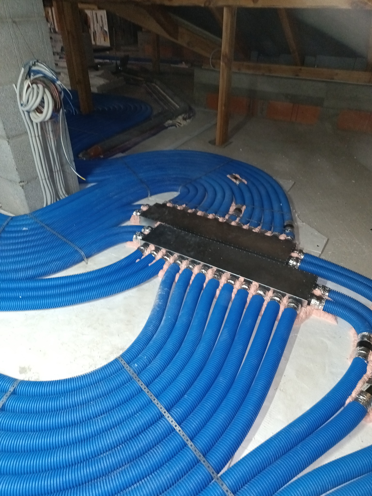 Niebieskie rury ogrzewania podłogowego, ułożone spiralnie i podłączone do rozdzielacza, widoczne w surowym stanie poddasza. Instalacja przed wylaniem posadzki.