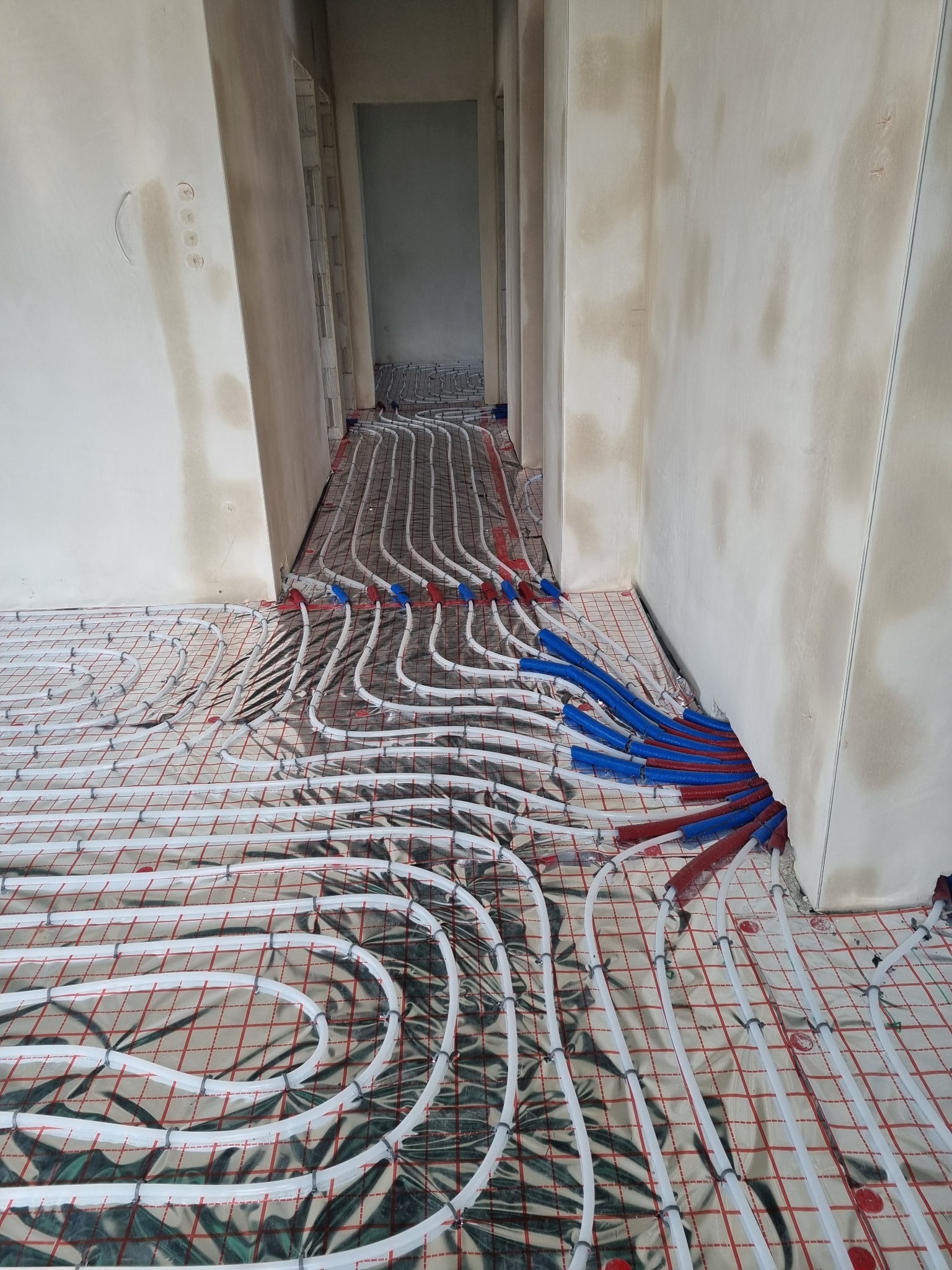 Instalacja ogrzewania podłogowego w trakcie montażu: białe rurki ułożone w pętle na folii z czerwoną siatką, nieotynkowane ściany w tle.