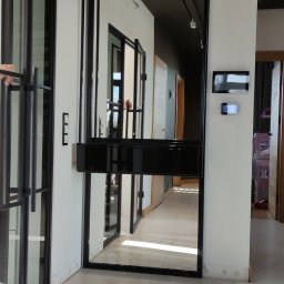 Cezary Stolarczyk Robione w metalu - Nowoczesne drzwi szklane z czarną ramą w długim korytarzu, widok z boku, minimalistyczny design.