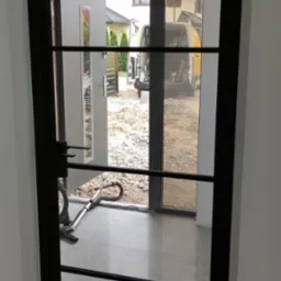 Nowoczesne drzwi z czarną ramą i szklanymi panelami, widok na plac budowy z zaparkowanym busem.