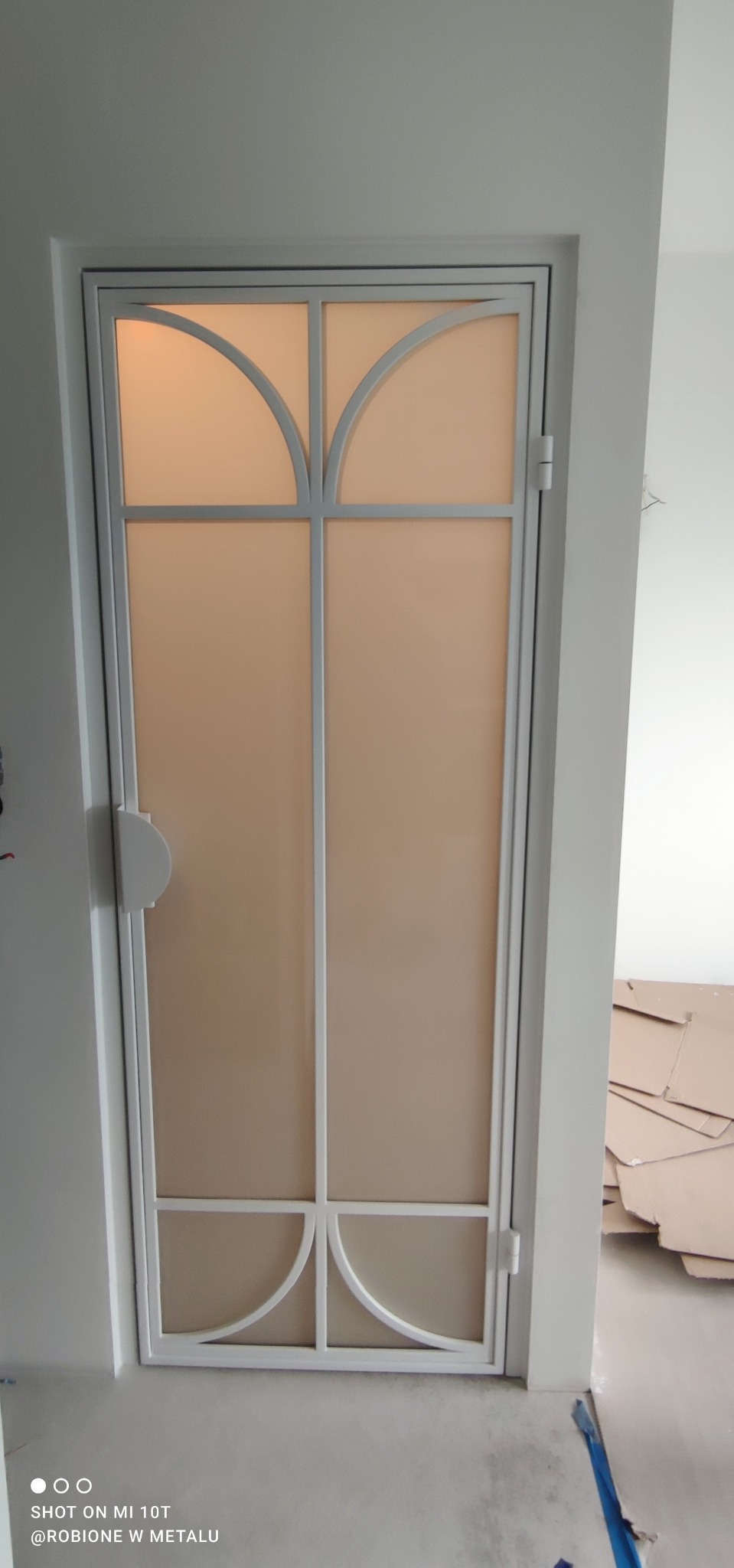 Zamknięte, białe drzwi wewnętrzne z matowego szkła, z metalową ramą o dekoracyjnych łukach w górnej i dolnej części, widoczne w trakcie remontu.