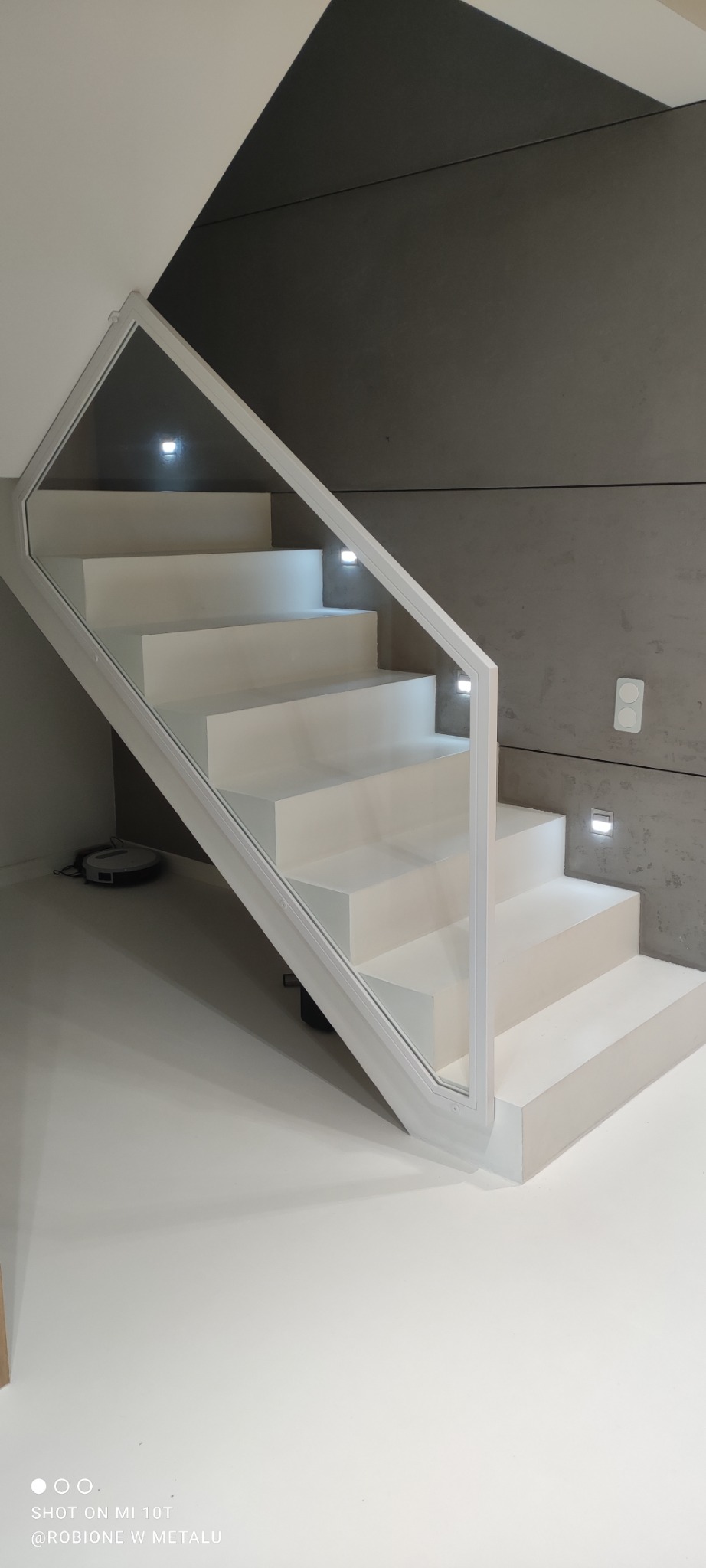 Nowoczesne, białe schody z podświetlanymi stopniami i szklaną balustradą, widok z dołu, na podłodze robot sprzątający.