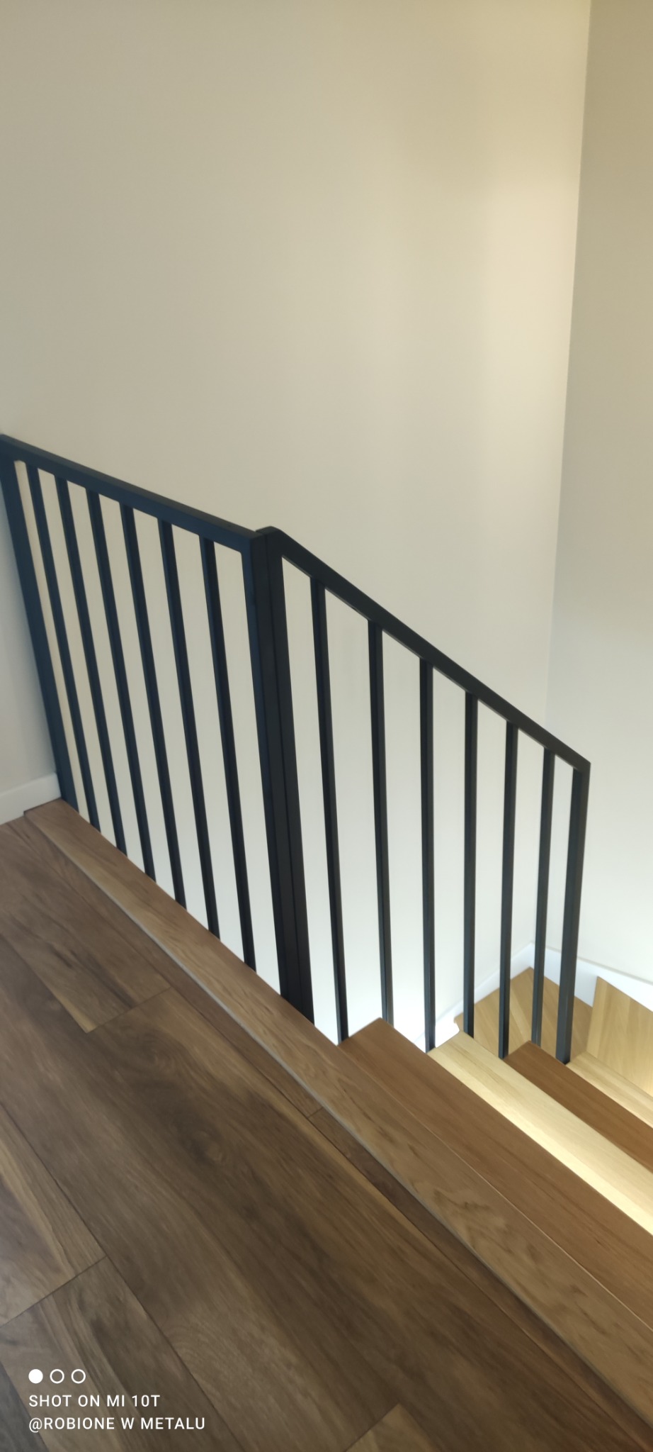 Nowoczesna, czarna poręcz schodowa z pionowymi szczebelkami na drewnianych schodach w jasnym wnętrzu.