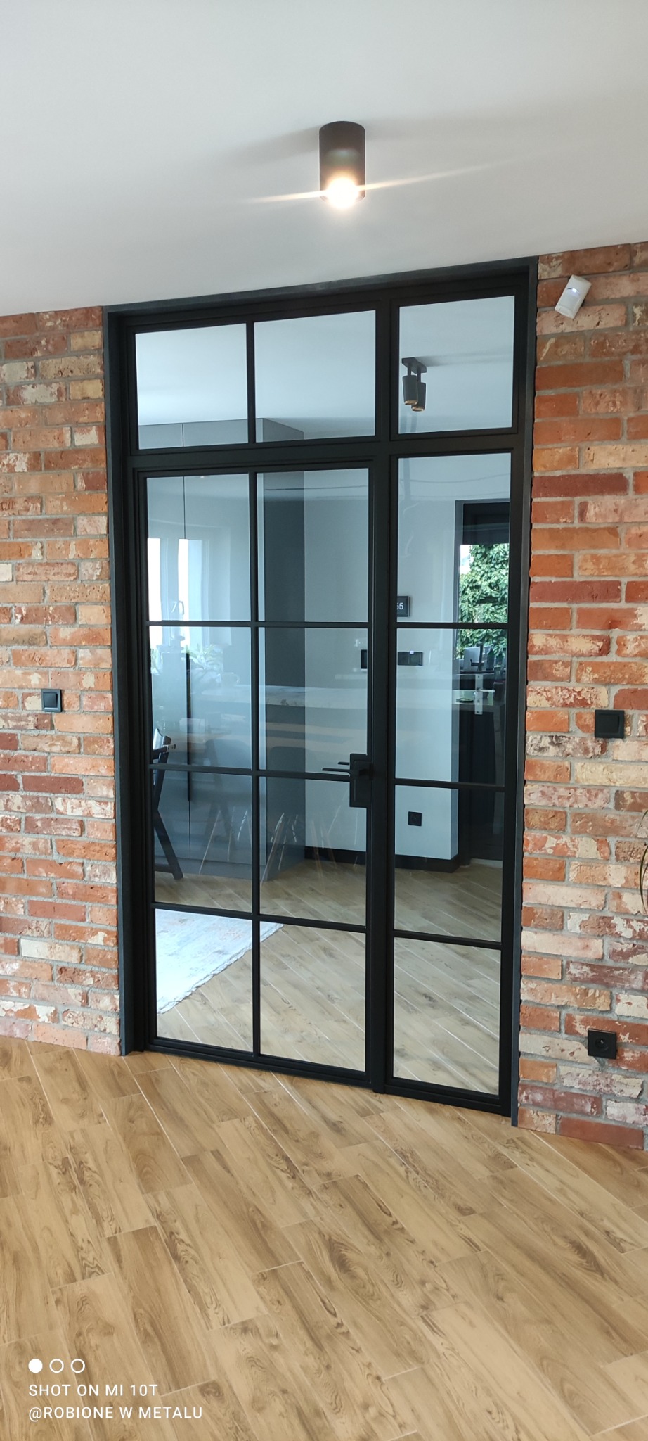 Dwuskrzydłowe drzwi z czarnymi szprosami, oddzielające pomieszczenia wykończone cegłą i drewnopodobnymi panelami, widok na jasne wnętrze.
