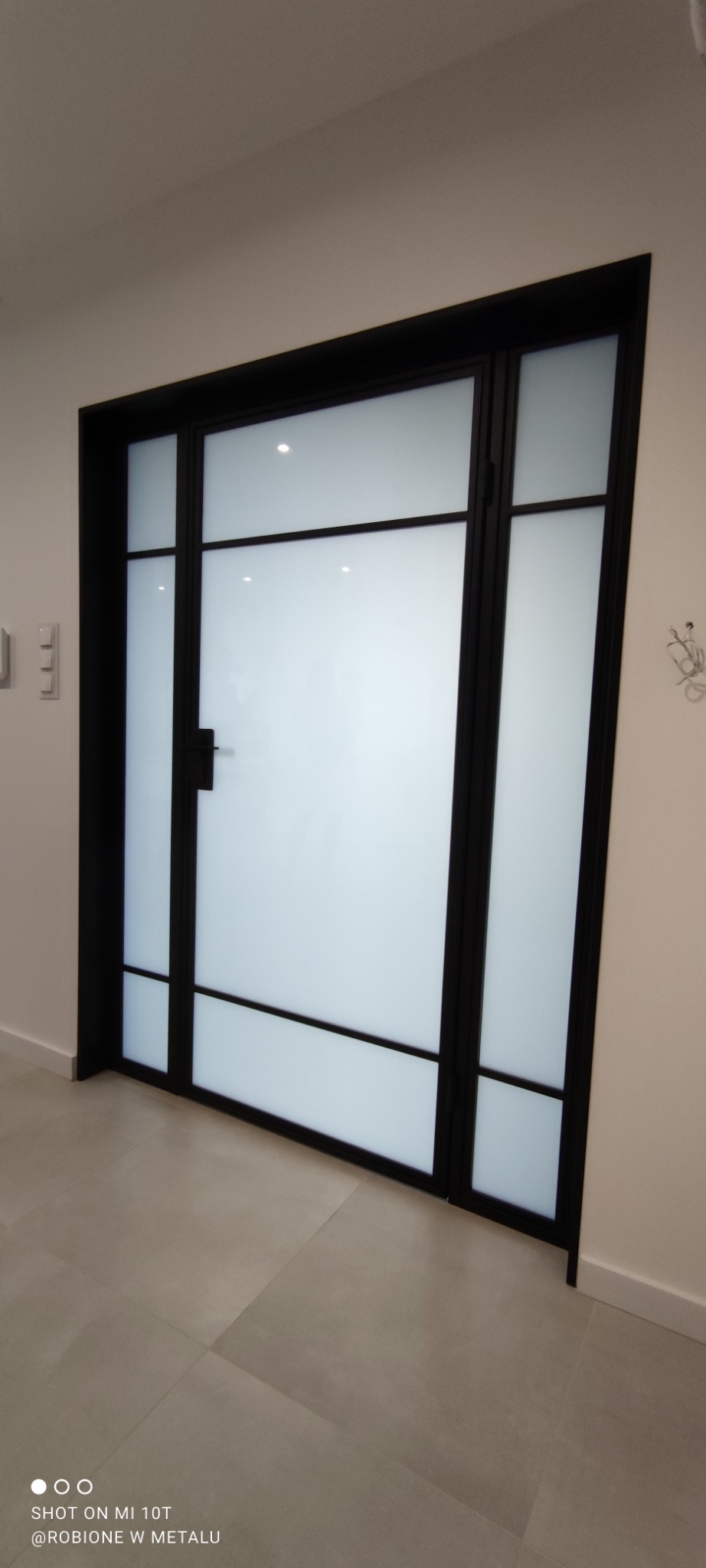 Nowoczesne drzwi wewnętrzne z czarną ramą i matowym szkłem, dzielone na prostokątne segmenty, widoczne na tle jasnej ściany i podłogi z płytek.