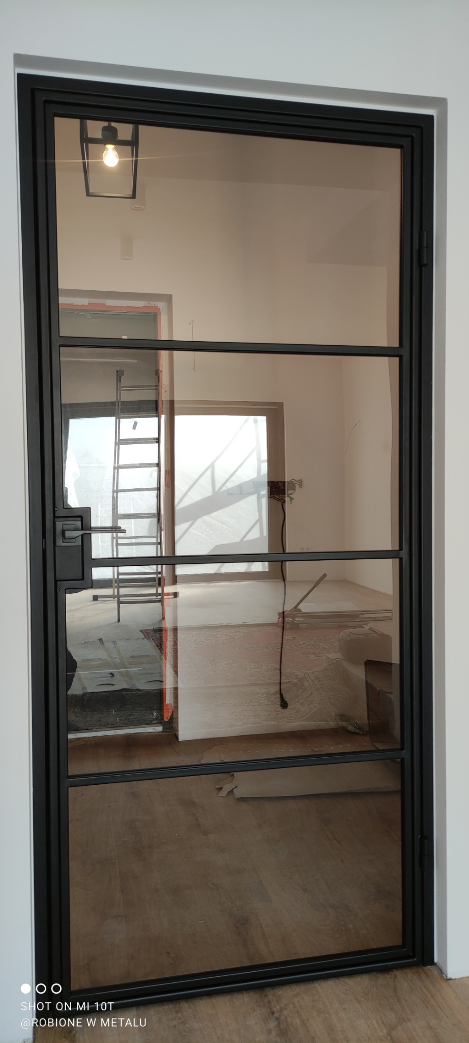 Czarna, szklana konstrukcja drzwi z metalowymi szprosami. W tle widoczne pomieszczenie w trakcie remontu, drabina oparta o ścianę i surowe podłoże.