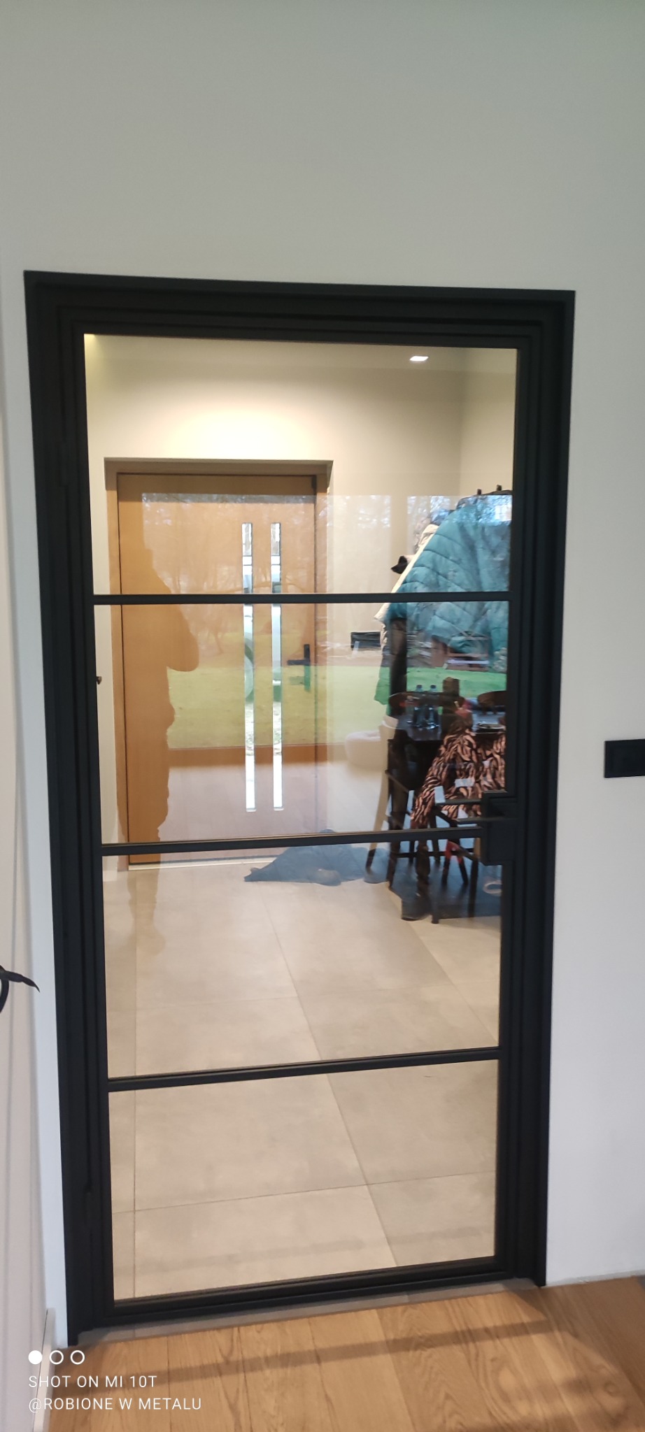 Szklane drzwi wewnętrzne z czarną, metalową ramą i trzema poziomymi szprosami, widoczne odbicie wnętrza domu