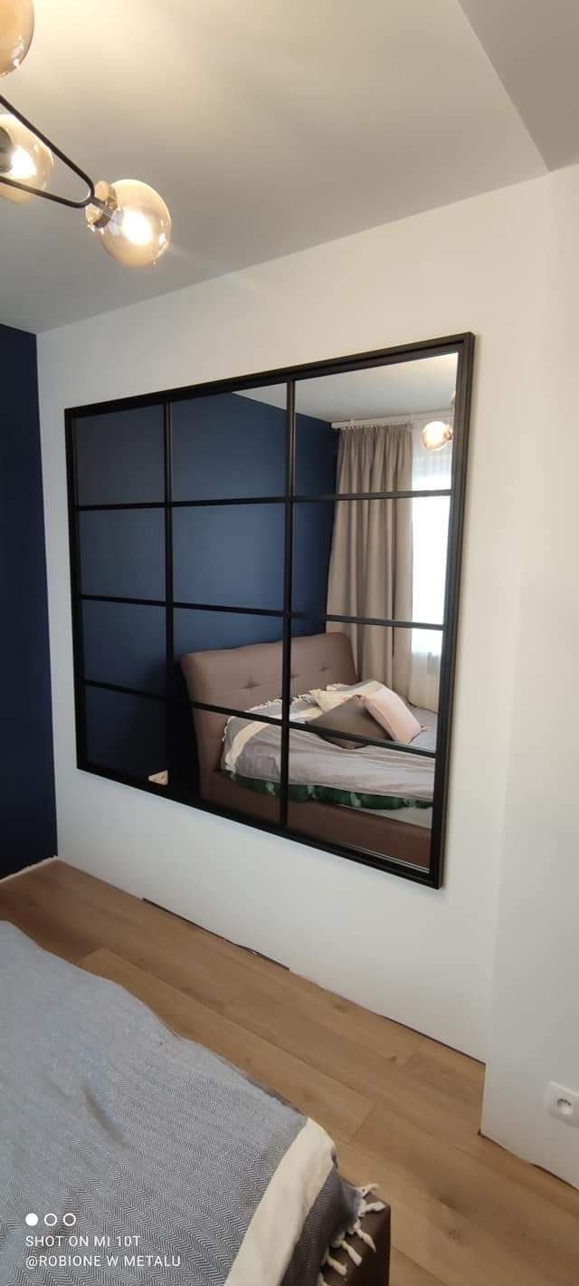 Czarna, stalowa konstrukcja z szybami, pełniąca funkcję okna wewnętrznego, oddzielająca sypialnię z łóżkiem i niebieską ścianą od reszty pomieszczenia, widoczna z perspektywy podłogi z jasnymi...