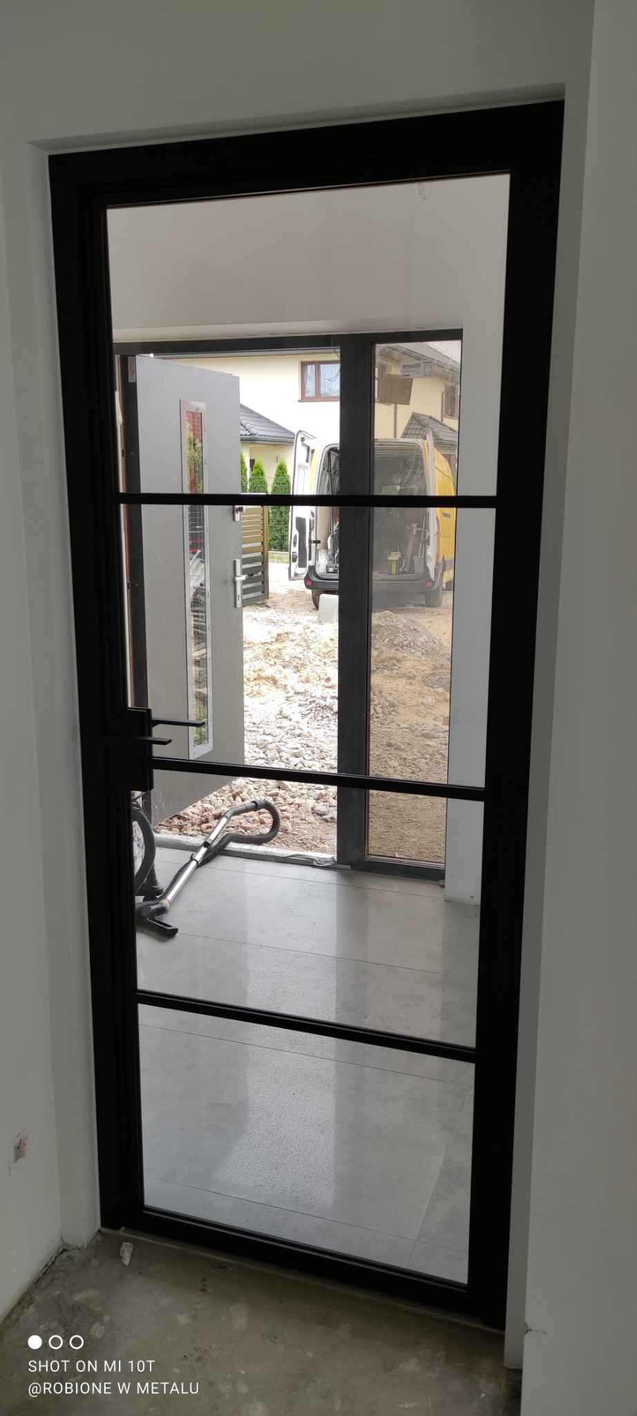 Nowoczesne drzwi z czarną ramą i szklanymi panelami, widok na plac budowy z zaparkowanym busem.