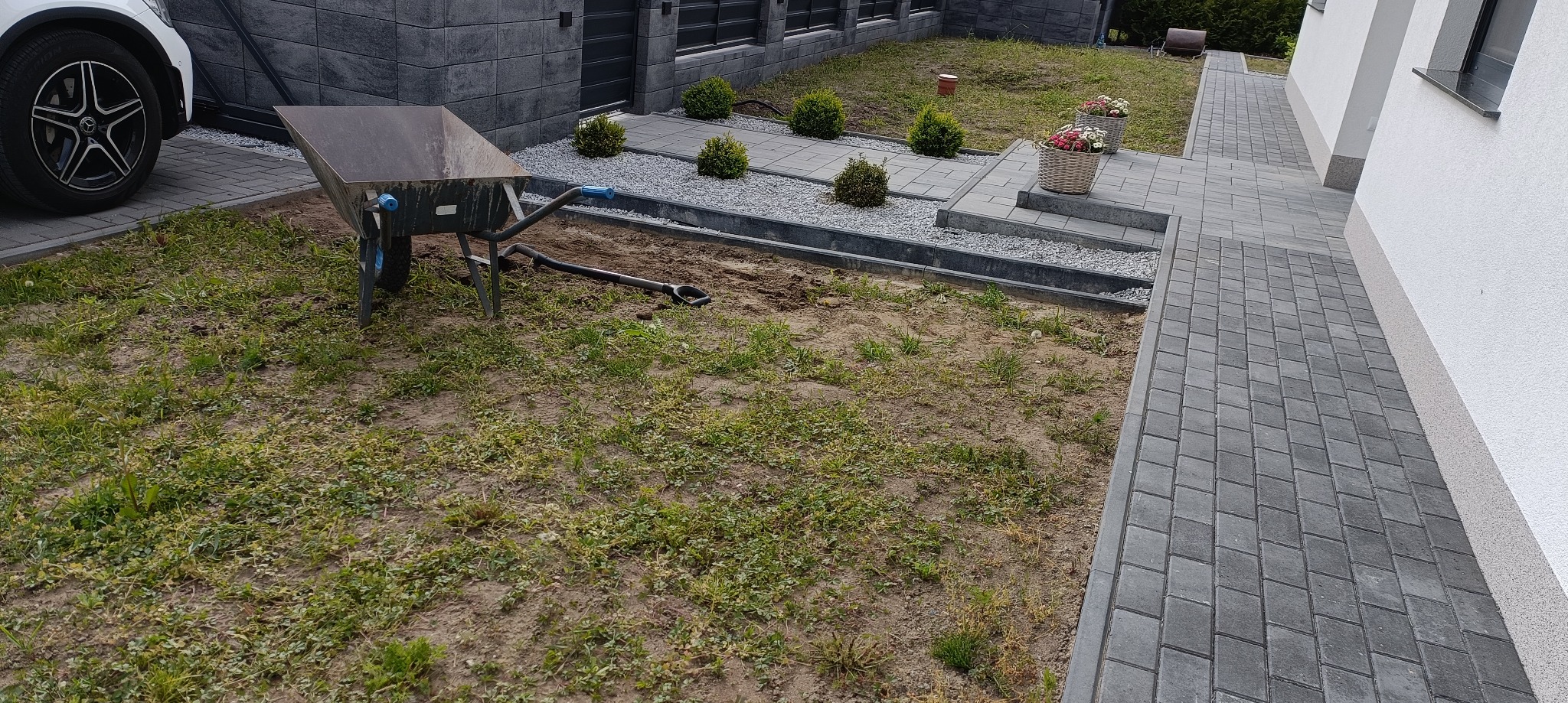Przygotowanie terenu pod trawnik: taczka, łopata i częściowo usunięta darń. W tle kostka brukowa, rabaty z krzewami i kwiatami. Mercedes na podjeździe.