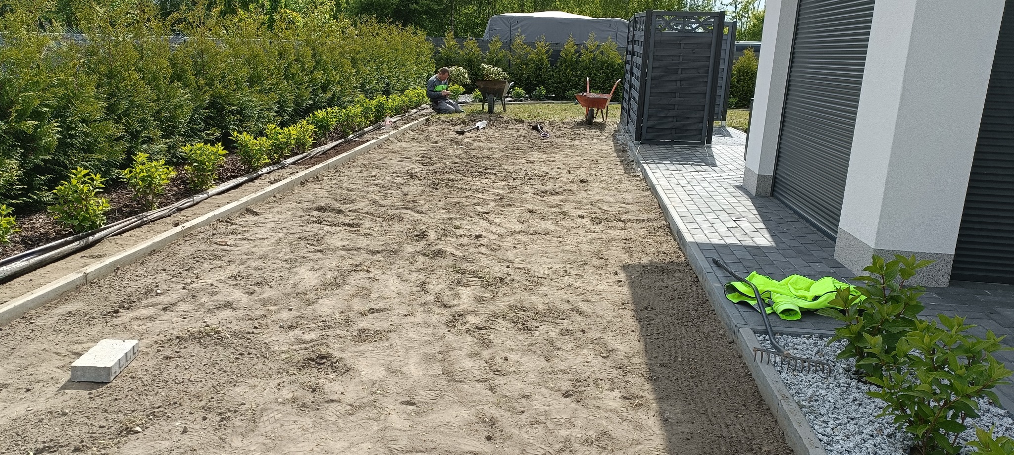 Przygotowany teren pod trawnik z rolki, z obrzeżami i systemem nawadniania. Pracownik z taczką w tle. Widoczny fragment budynku i roślinność.