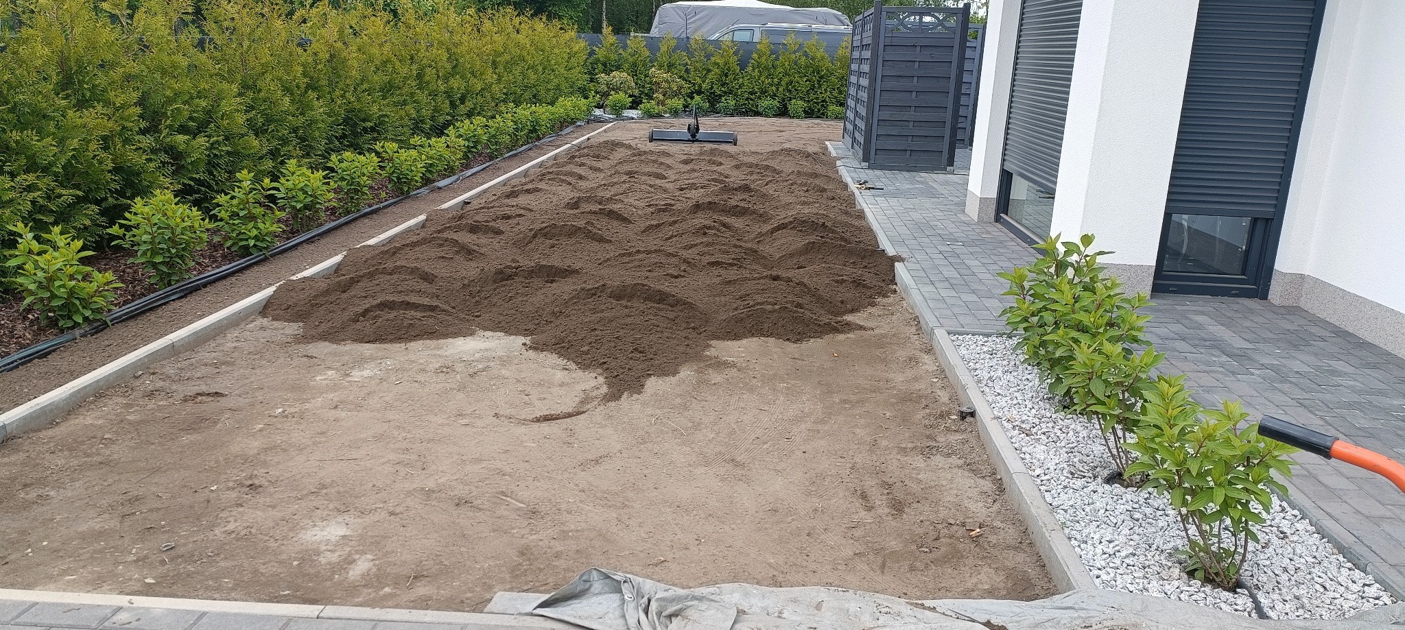 Przygotowanie podłoża pod trawnik: wyrównana ziemia w ogrodzie, obrzeża z kostki, rośliny wzdłuż ścieżki. Widoczny wał do trawy i fragment instalacji nawadniającej.