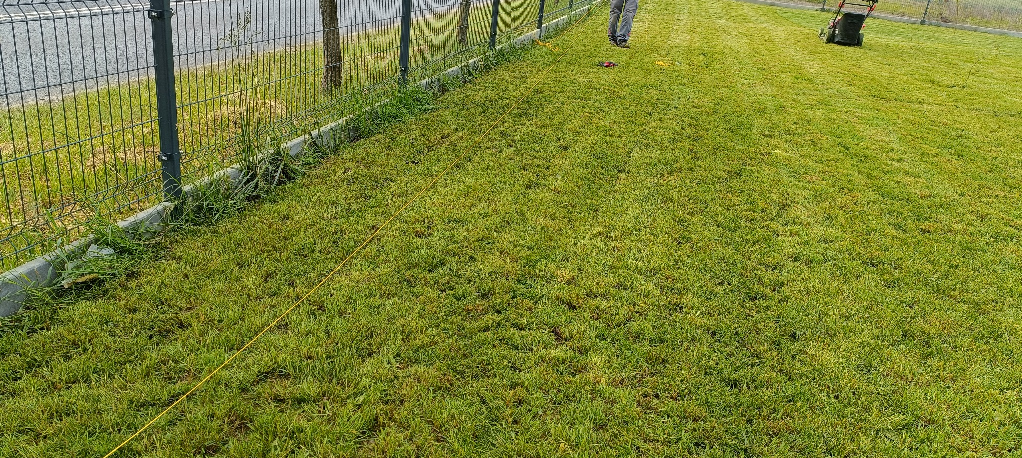 Świeżo skoszona trawa z rolki wzdłuż ogrodzenia, widoczna linia wyznaczająca obszar cięcia i fragment kosiarki na trawniku. Częściowo widoczna osoba nadzorująca prace.